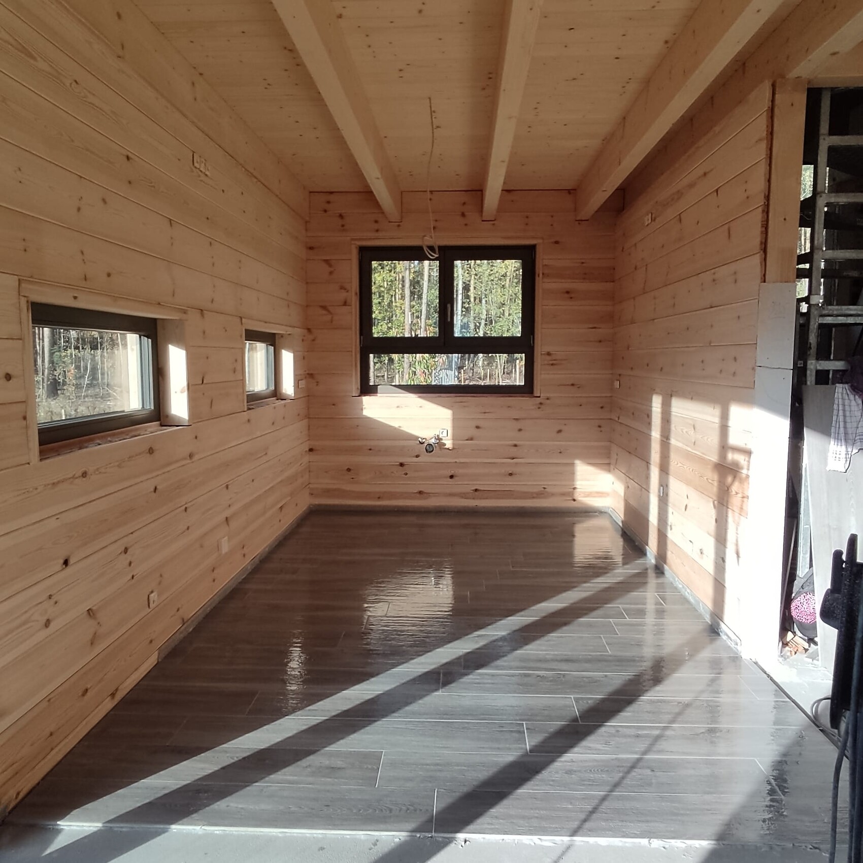 Heller Innenraum aus Holz im Bau: mehrere Fenster, glänzender grauer Boden, Bauwerkzeuge an der Wand.