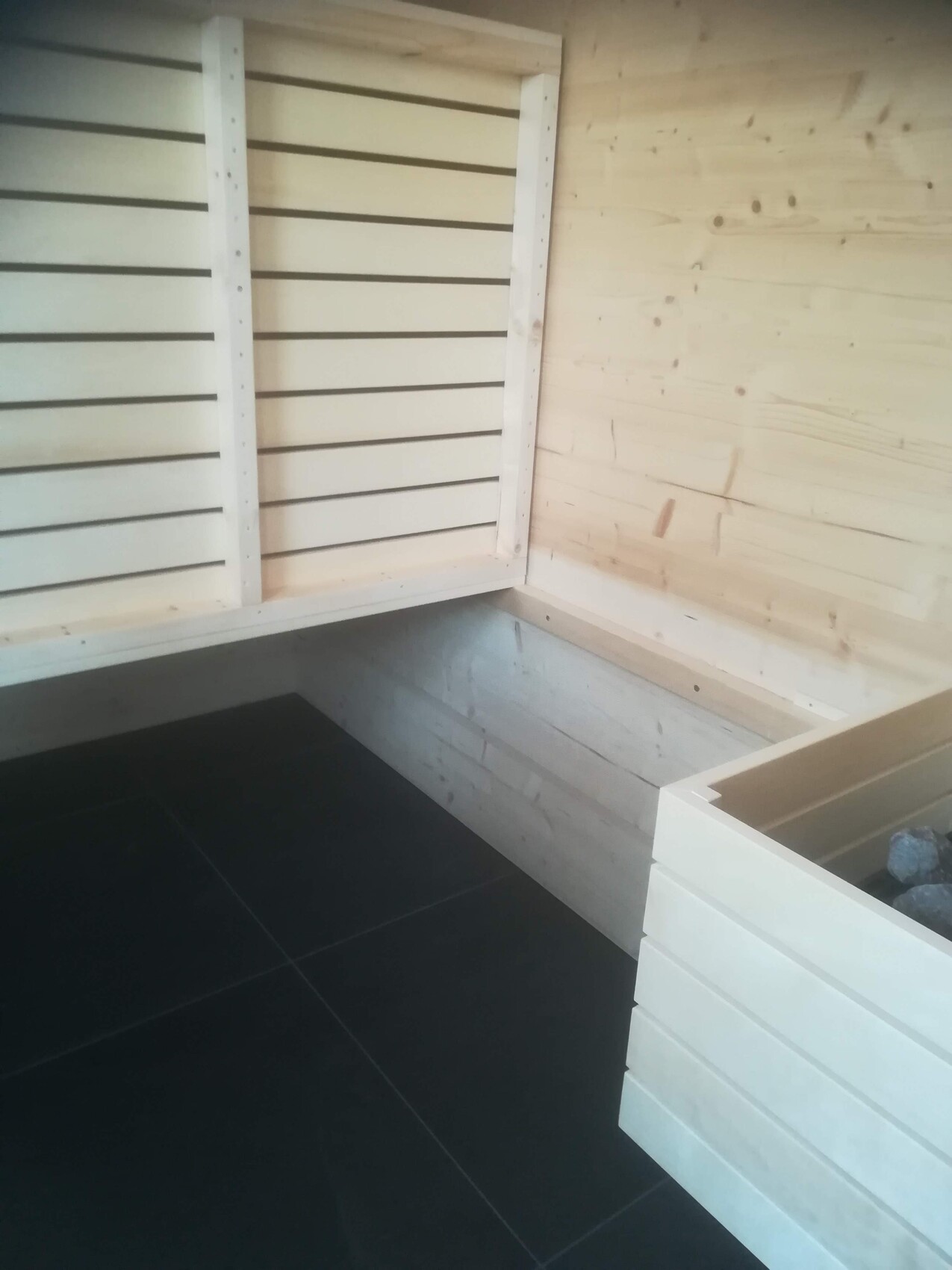 Sauna-Innenraum aus Holz mit Eckbank, dunklem Boden und Heizbereich mit Steinen.
