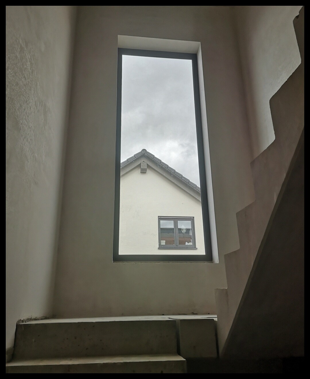 Blick durch ein schmales Innenfenster auf den Dachgiebel eines Nachbarhauses bei grauem Himmel.