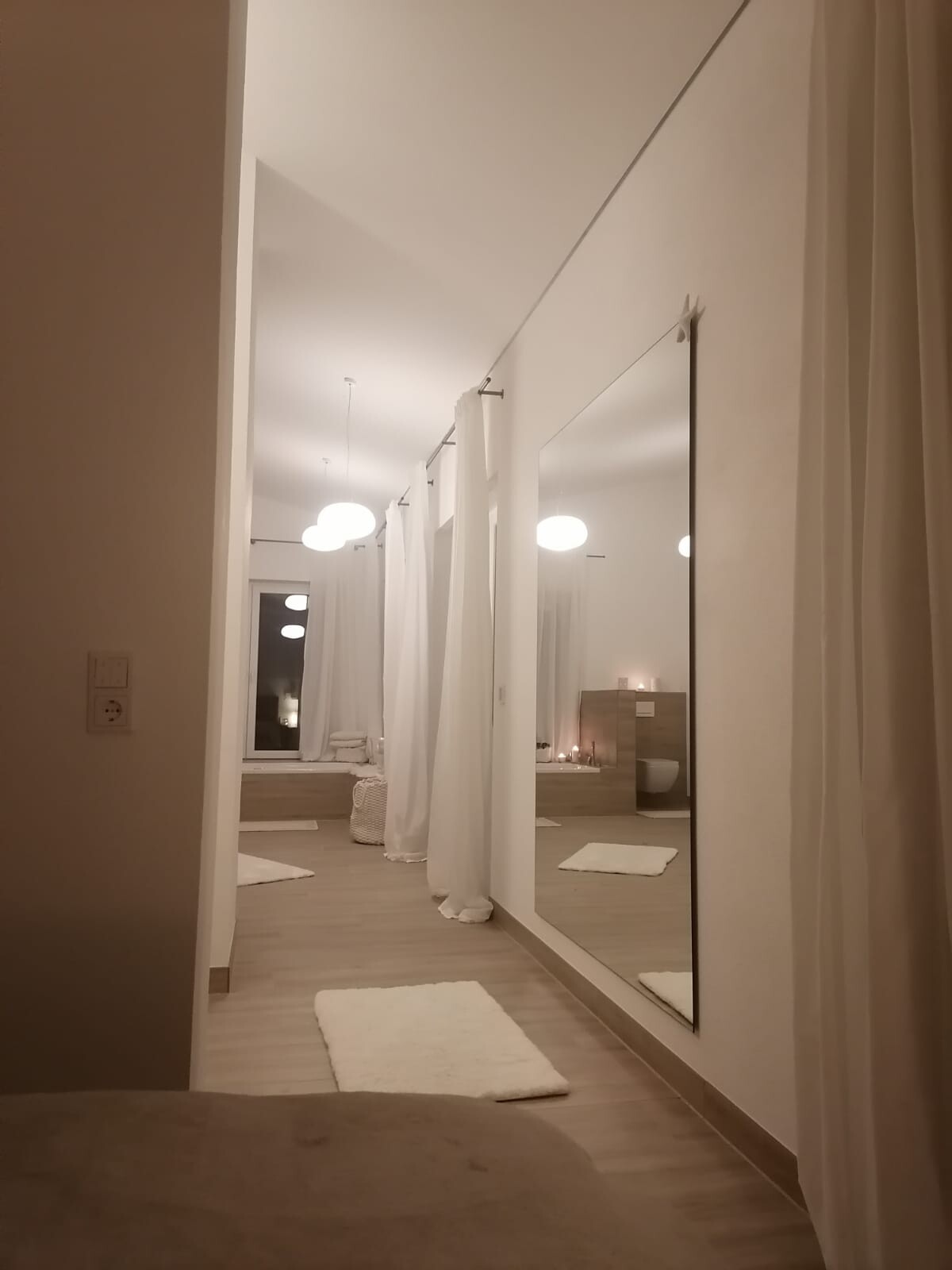 Langer heller Behandlungs-/Spa-Raum mit weißen Vorhängen, Holzfußboden und Spiegelwand.