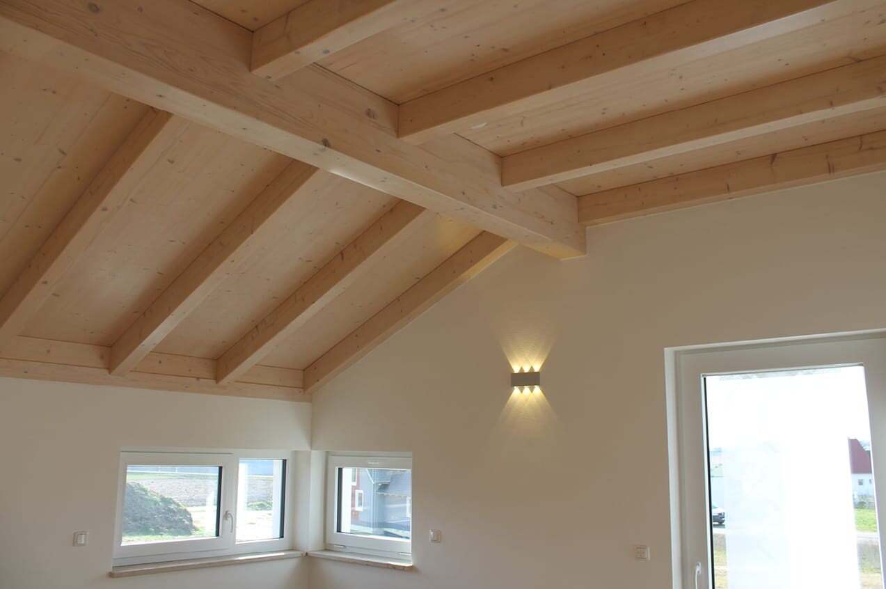 Innenraum mit sichtbarer Holzbalkendecke, weißen Wänden, Wandlampe und Fenstern.
