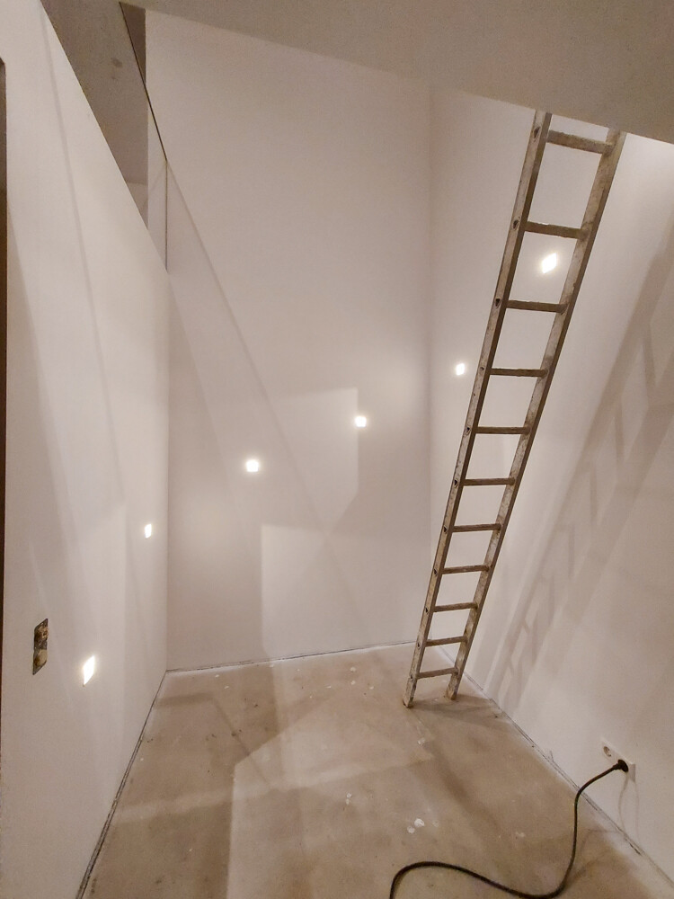 Leerer Bauraum mit Leiter an der Wand, Wandbeleuchtung sichtbar, Kabel auf dem Boden.