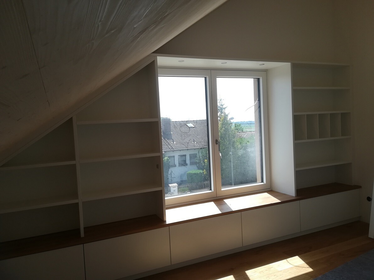 Heller Raum mit Dachschräge, weiße Regale um großes Fenster mit Fensterbank und Unterschränken.