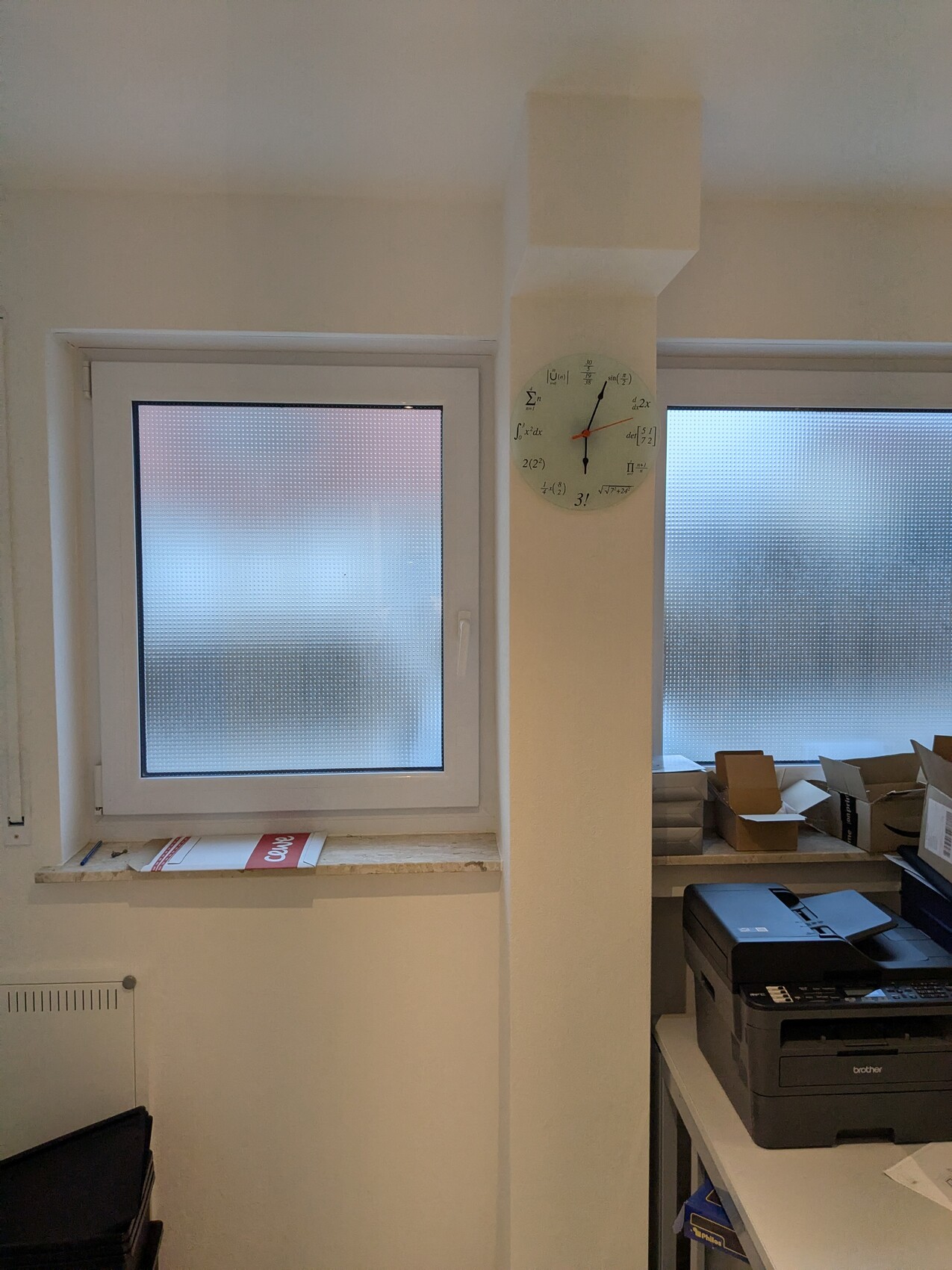 Büro mit zwei milchigen Fenstern, zentrale Säule mit Uhr aus Formelsymbolen, Drucker und Kartons.