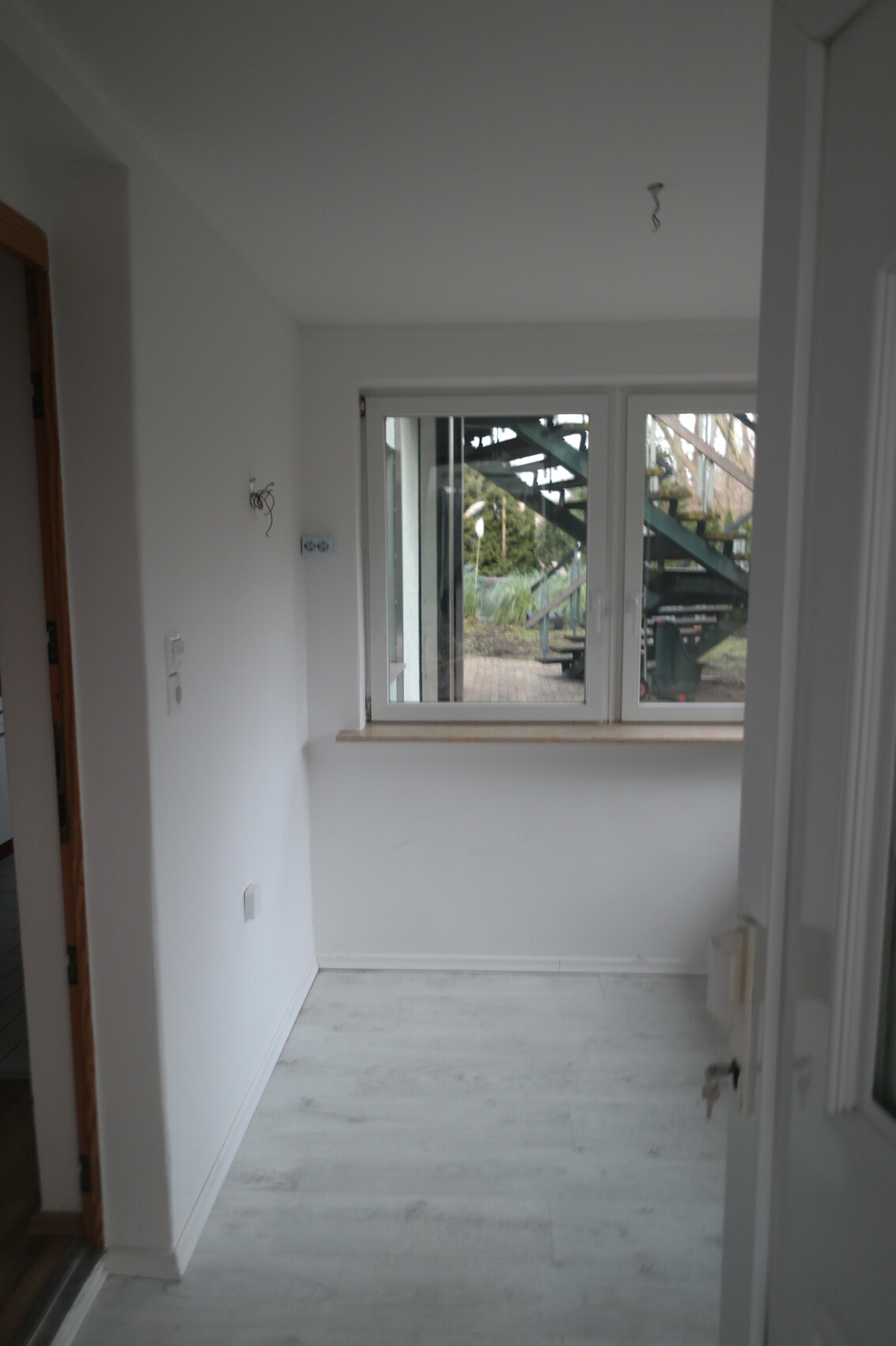 Heller, leerer Innenraum mit Fenster zur Außentreppe; freiliegende Kabel an der Wand.