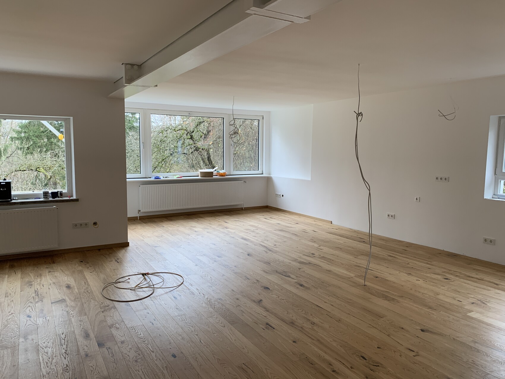 Leerer Raum mit Holzboden, großen Fenstern, Heizkörpern und herabhängenden Kabeln.