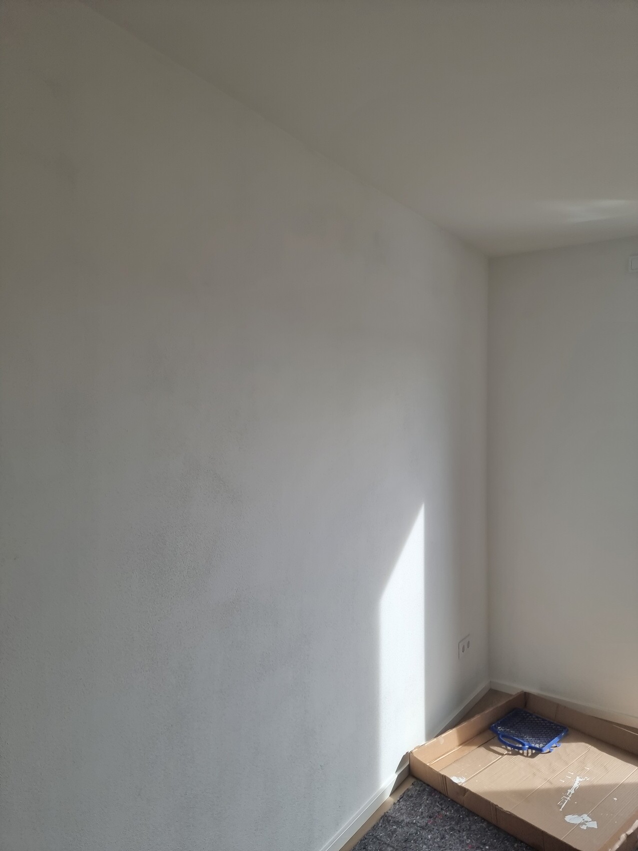 Weißes, leeres Zimmereck mit gestrichenen Wänden, Lichtfleck an der Wand, Kartonboden mit blauem Ding.