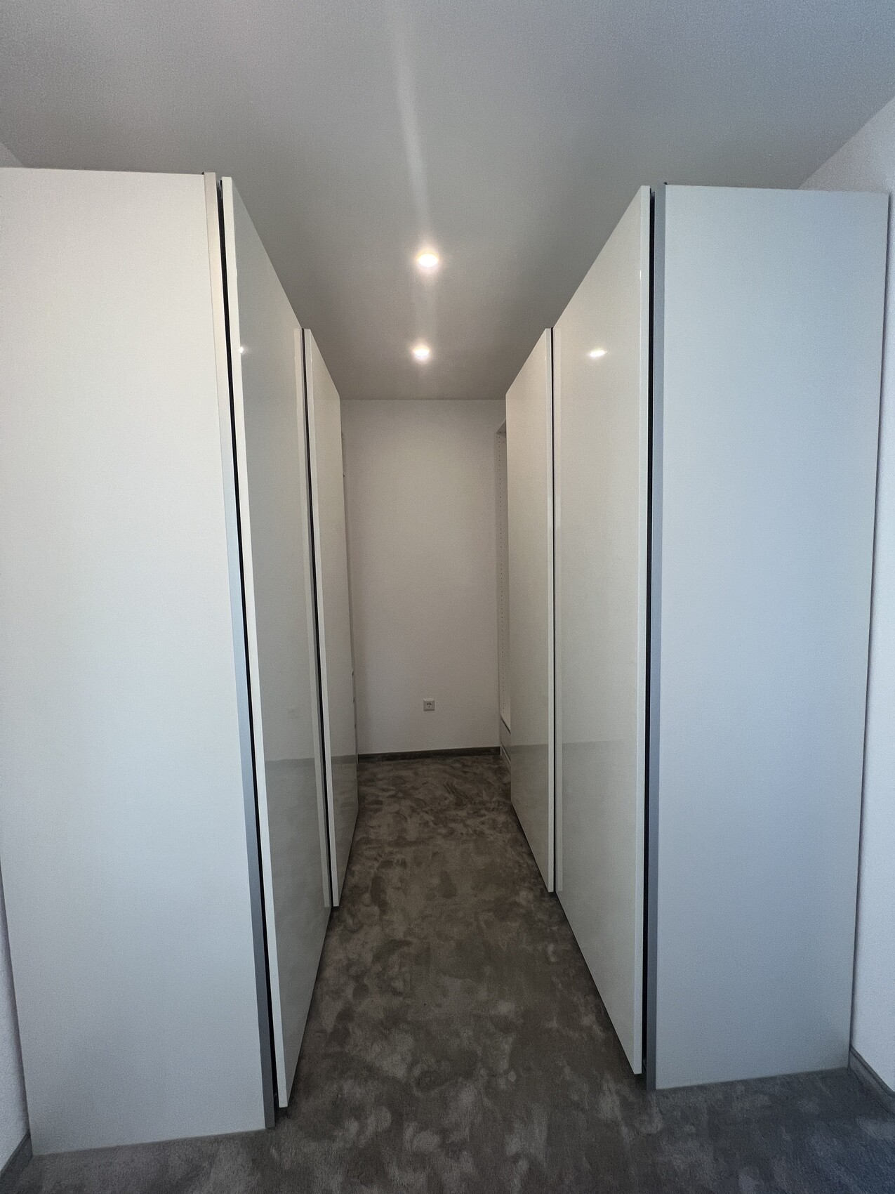 Begehbarer Kleiderschrank-Gang mit weißen Schränken links und rechts, grauer Boden, Deckenleuchten