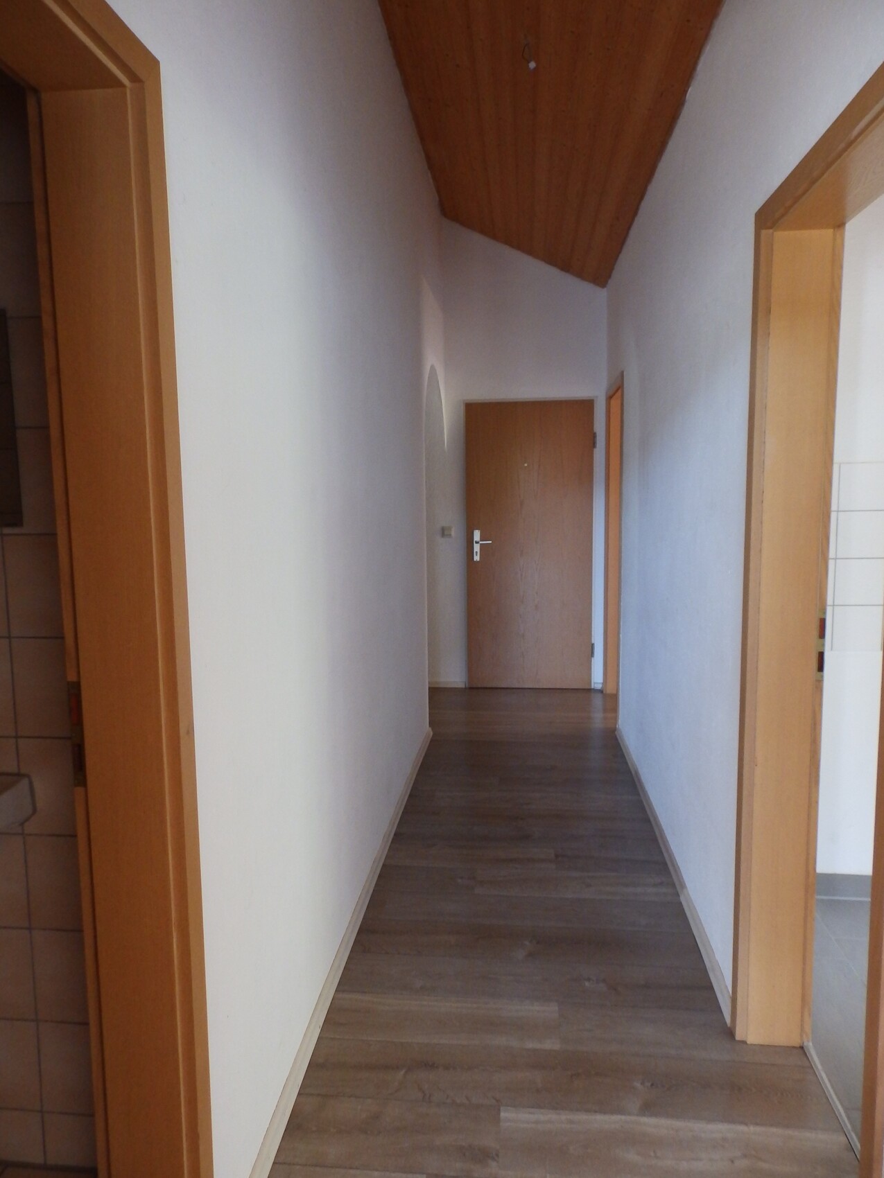 Langer Flur in einem Haus: weiße Wände, Holztüren, Holzfußboden, schräg verlaufende Holzdecke, Tür am Ende.