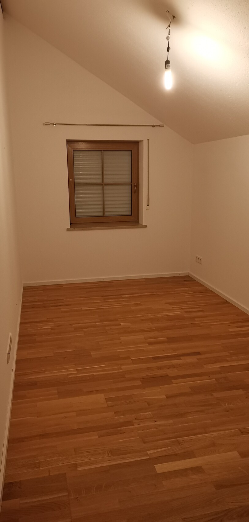Helles leeres Dachgeschosszimmer mit Holzboden, Holzfenster mit Rollladen, Glühbirne an der Decke.