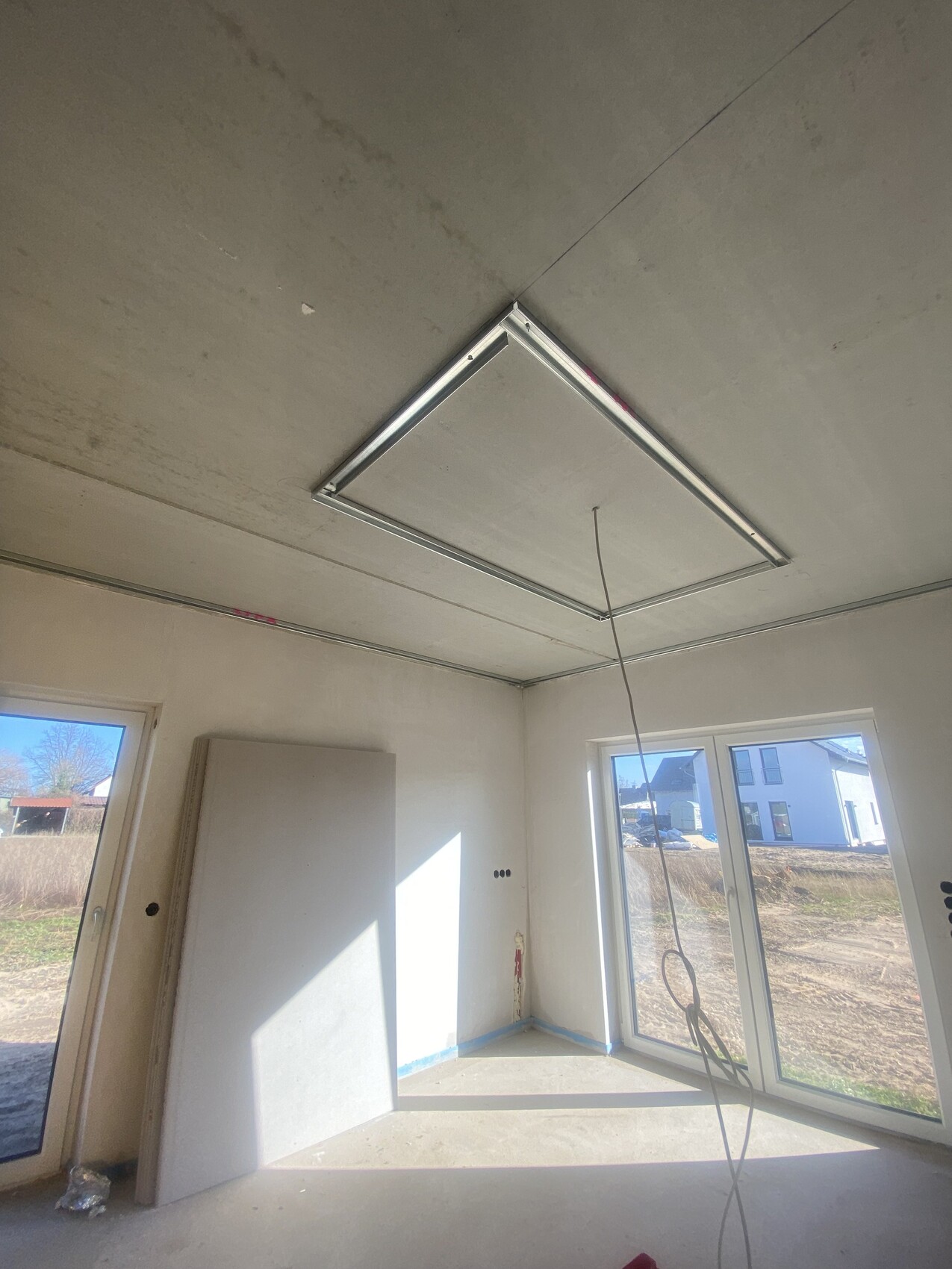 Unfertiger Innenraum: quadratischer Deckeneinbau-Rahmen hängt, Kabel fällt herab, große Fenster.
