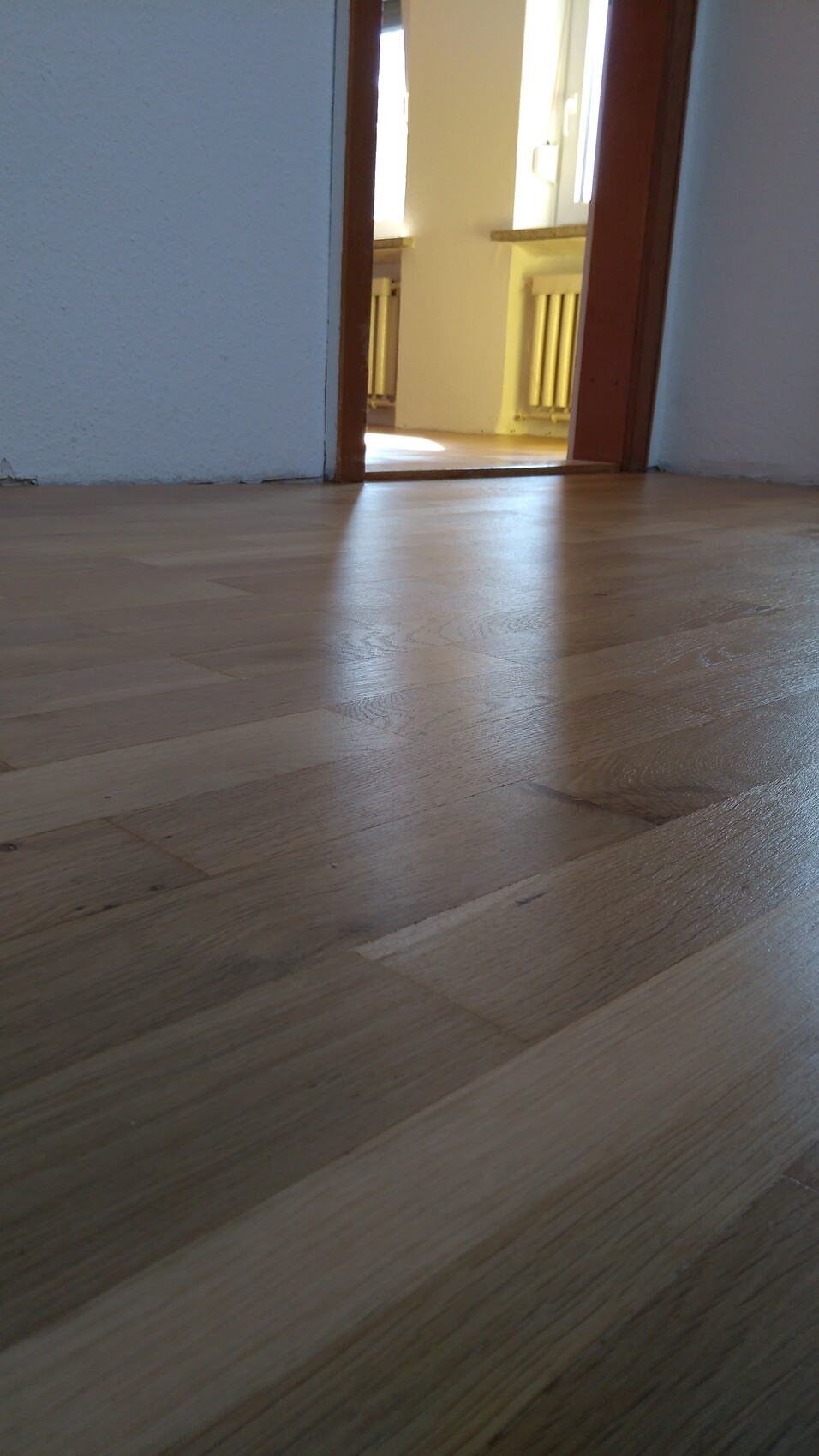 Blick auf Holzboden im Vordergrund, offene Tür führt zu hellem Raum mit Heizkörpern.