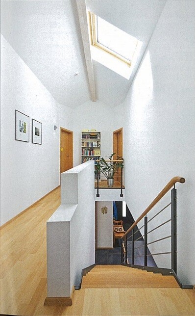Heller Innenraum mit Treppe, Geländer, Bücherregal und Pflanze am Ende eines Flurs.