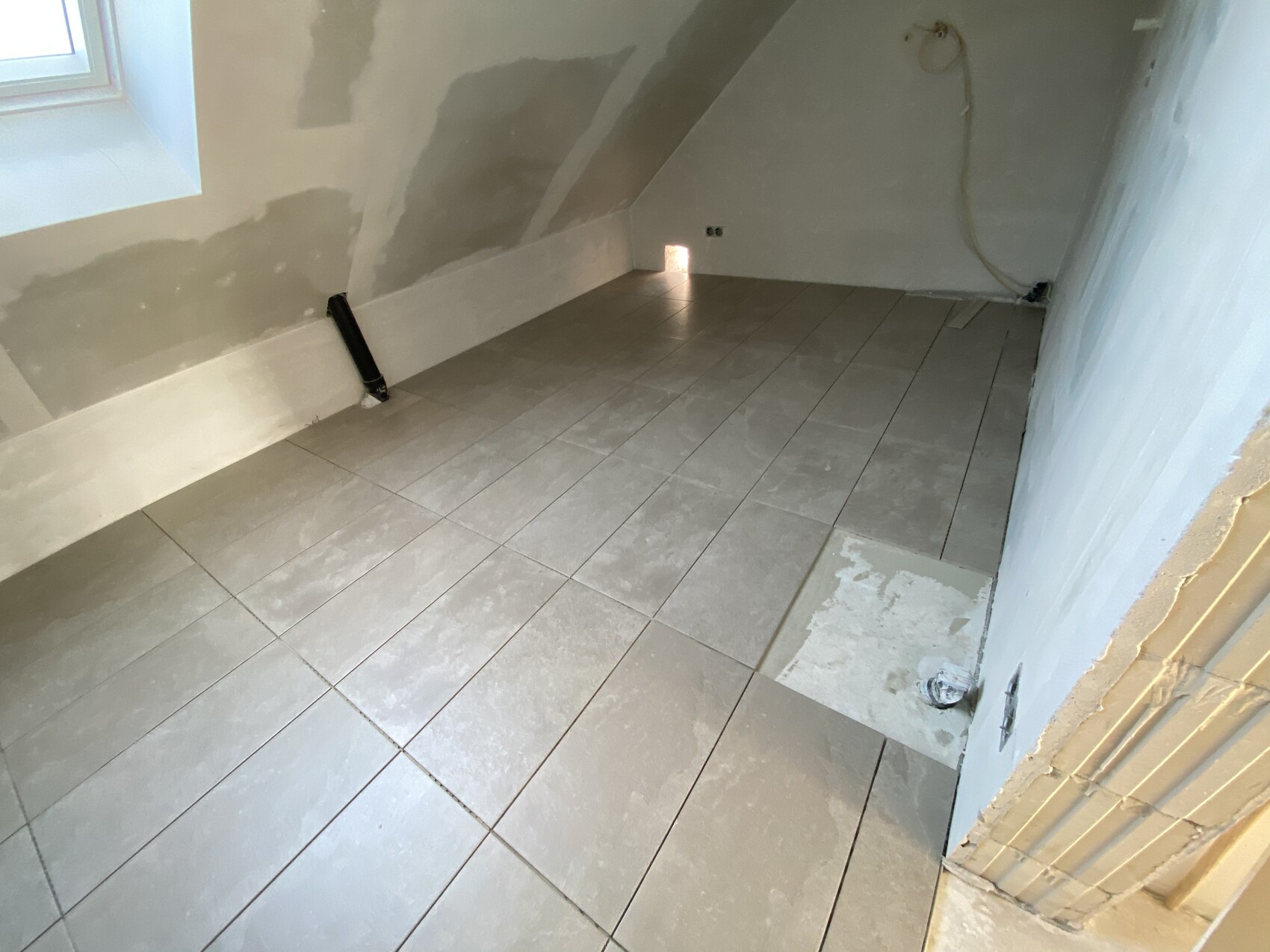 Kellerraum mit grauen Bodenfliesen, Dachschräge, unfertigen Wänden und sichtbaren Leitungen.