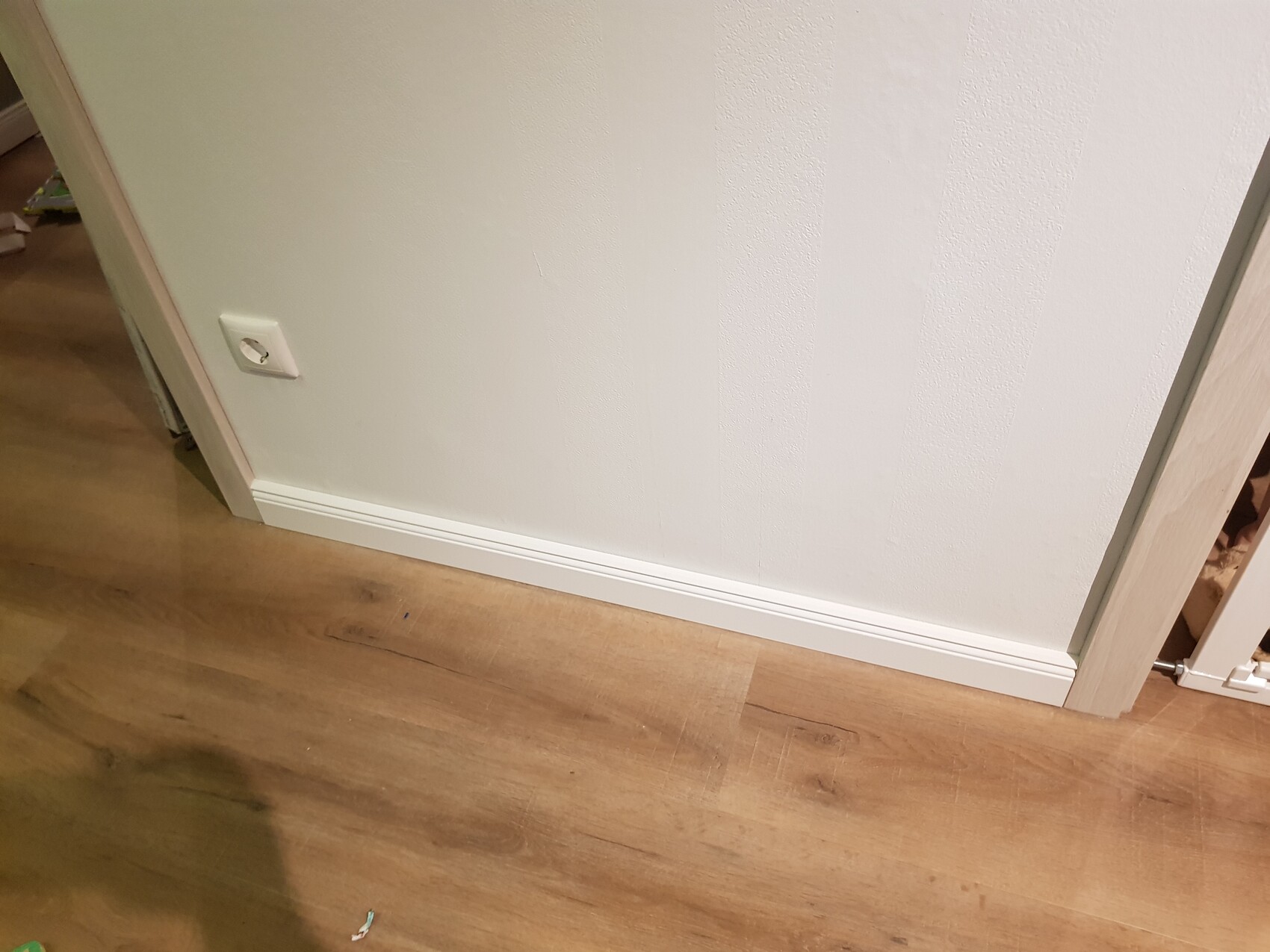 Weiße, texturierte Innenwand mit Sockelleiste; Steckdose links am Boden, Holzboden.