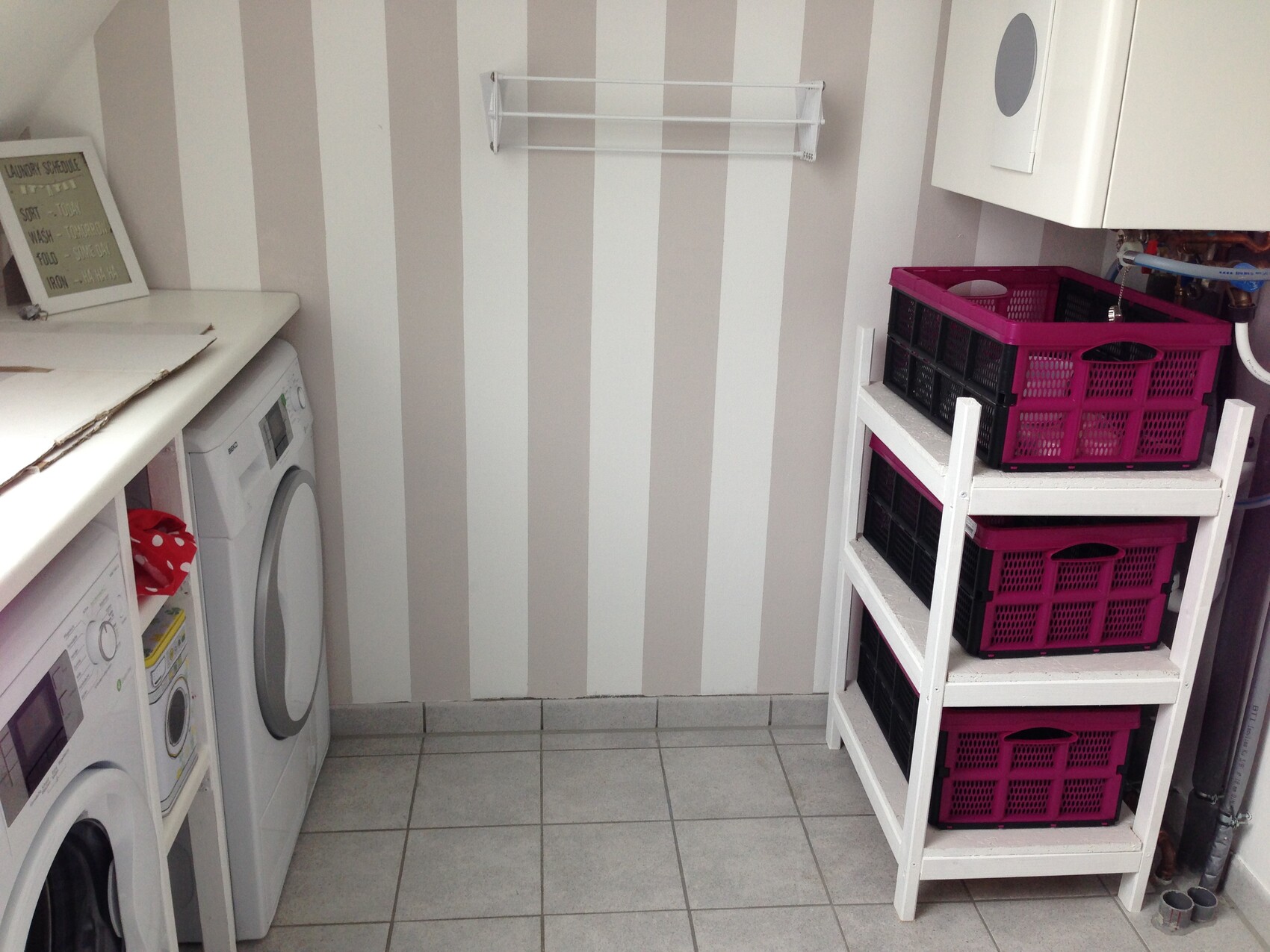 Waschraum mit Frontlader-Waschmaschinen links, weiße Regale mit pinken Körben rechts, gestreifte Wand.