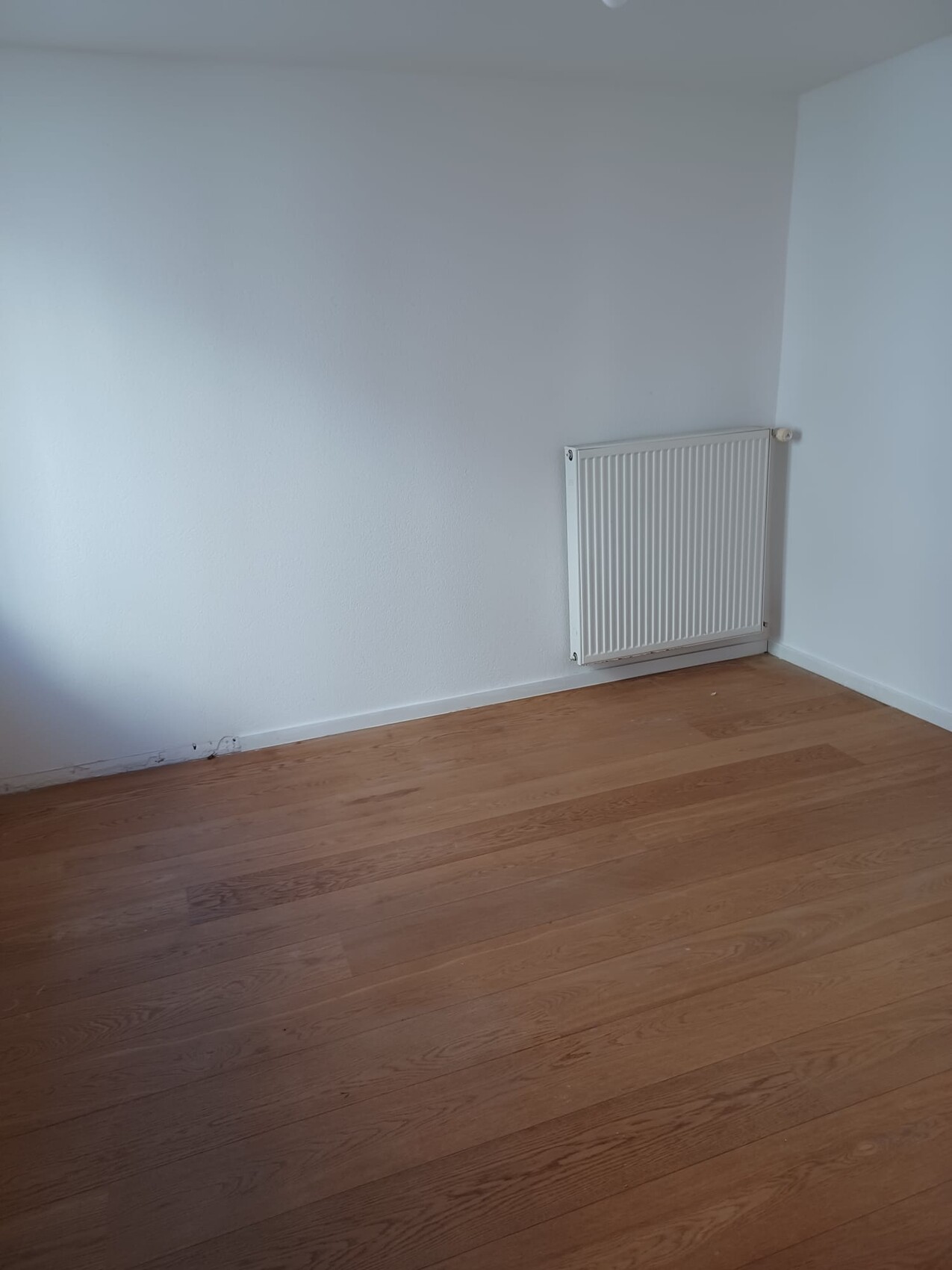 Leerer Raum mit Holzfußboden, weißen Wänden und Heizkörper rechts an der Wand.