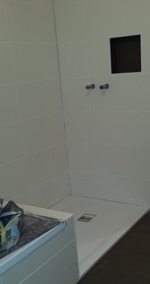 Dusche mit weißen Fliesen, zwei Armaturen (rot/blau), Wandnische; links Waschmaschine.