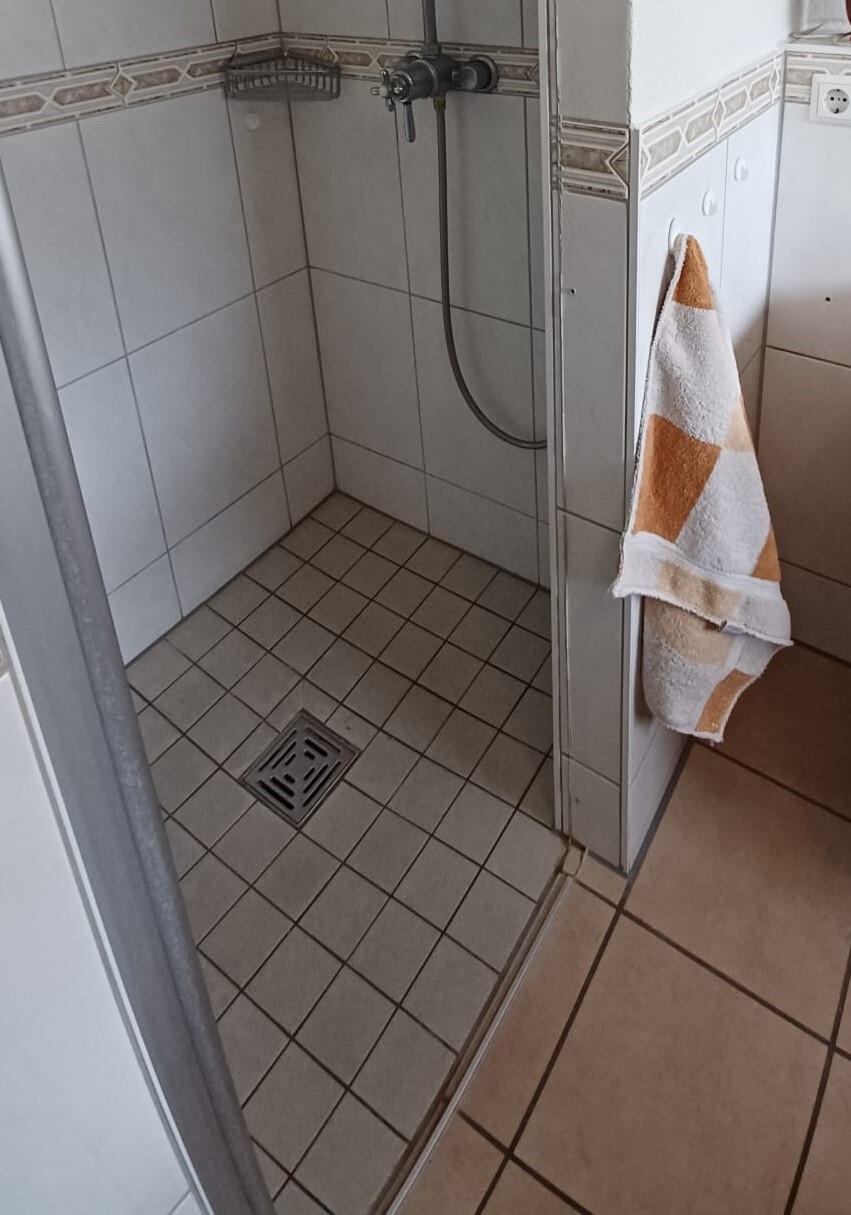 Kleines Duschbad mit Wand-Handbrause, Fliesenboden, Bodenabfluss, orange-weiß gemustertes Handtuch.