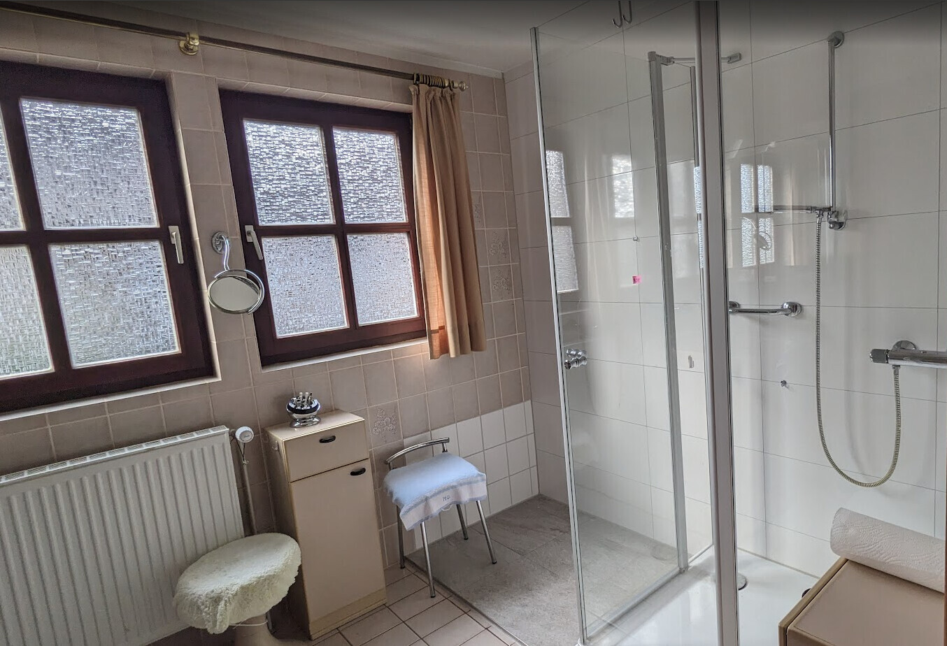 Badezimmer mit Glasdusche, zwei gefrostete Fenster, Hocker, schmaler Waschschrank und Spiegel.
