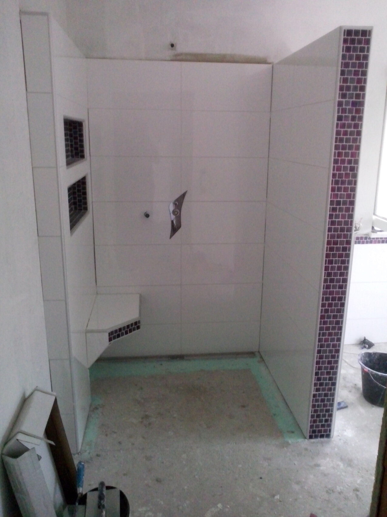 Duschbereich unter Bau mit weißen Fliesen, dunkler Mosaiknische und Sitzbank.