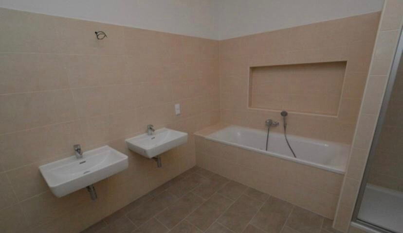 Badezimmer mit zwei Wandwaschbecken, Badewanne mit Nische, beige Fliesen und Glasdusche.