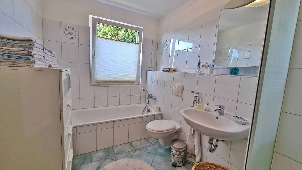 Helles Badezimmer mit Badewanne, Waschbecken, Toilette, Fenster mit Jalousie und Handtuchstapeln.