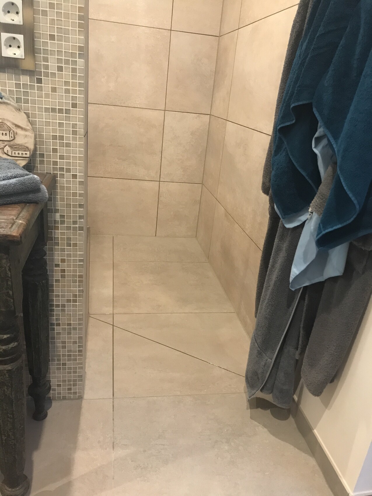 Kleines Badezimmer mit beigen Fliesen, Mosaikleiste links, Handtücher rechts, Bank links.