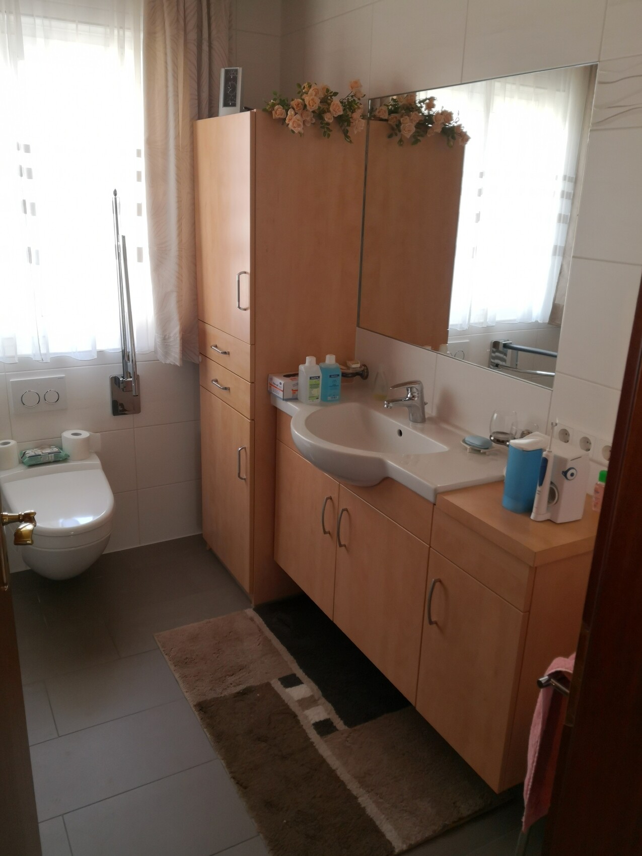 Badezimmer: Waschtisch mit Holzschränken, Spiegel, Blumen auf dem Schrank, Toilette links, Fenster.