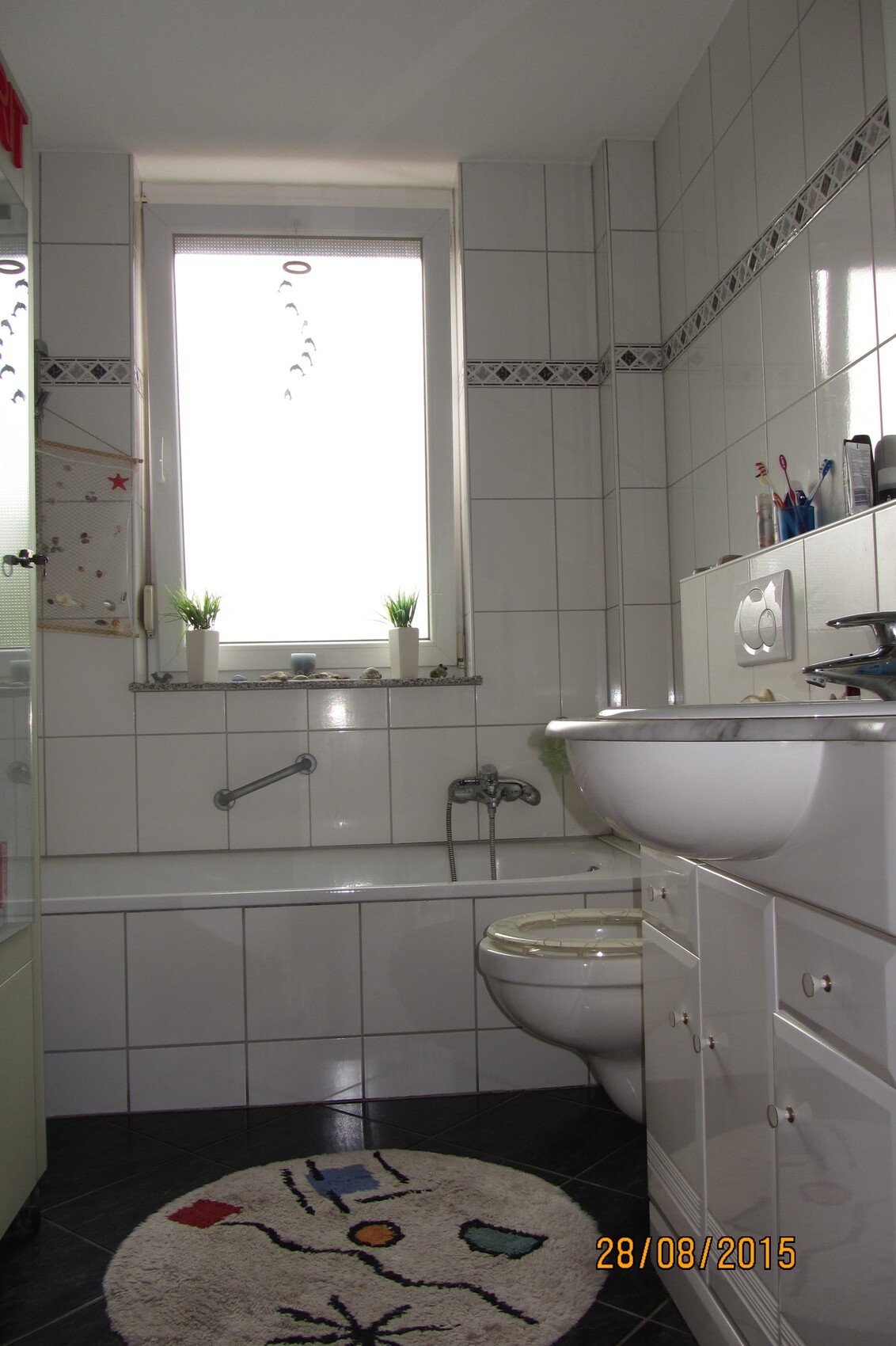 Helles Badezimmer mit Badewanne, WC, Waschbecken, Fenster und dekorativem Teppich.