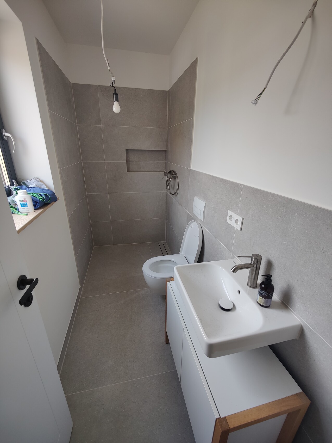 Unfertiges Badezimmer mit Toilette, Waschbecken, grauen Fliesen und Dusche-Nische.