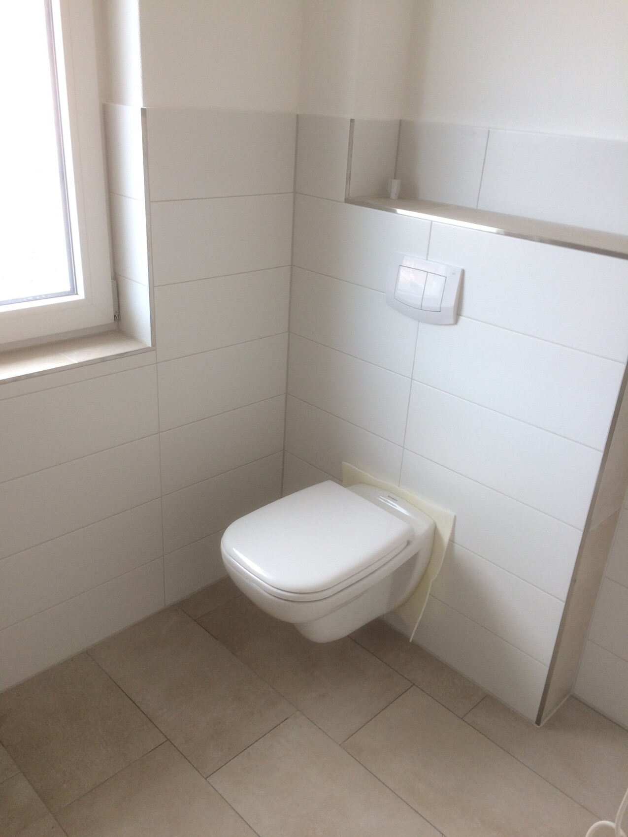 Wandhängendes WC mit Spülplatte in hellem Badezimmer mit weißen Fliesen und Fenster.