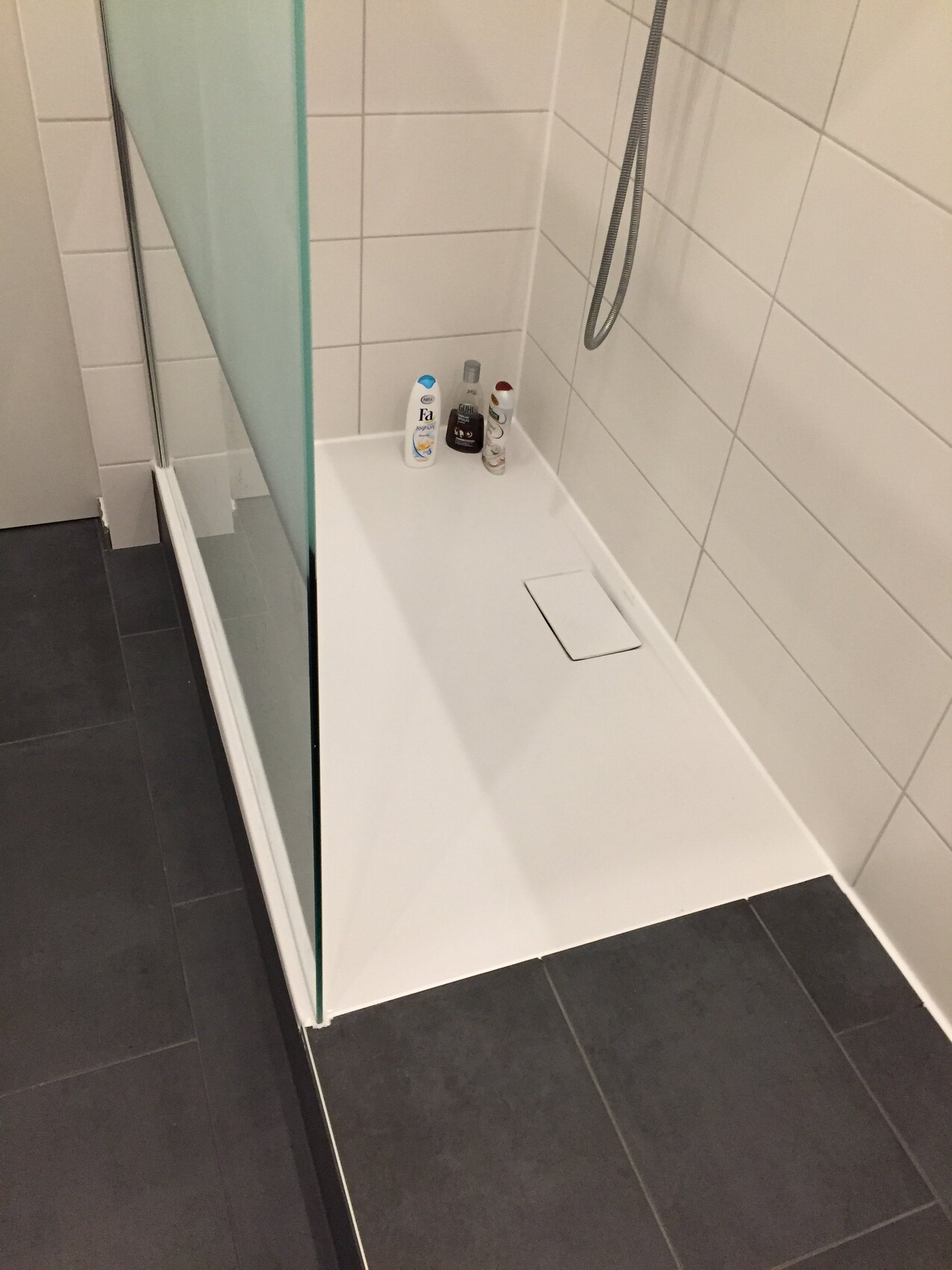 Helles Badezimmer mit Glasduschabtrennung, weißen Fliesen und Dusche; Badutensilien am Rand.