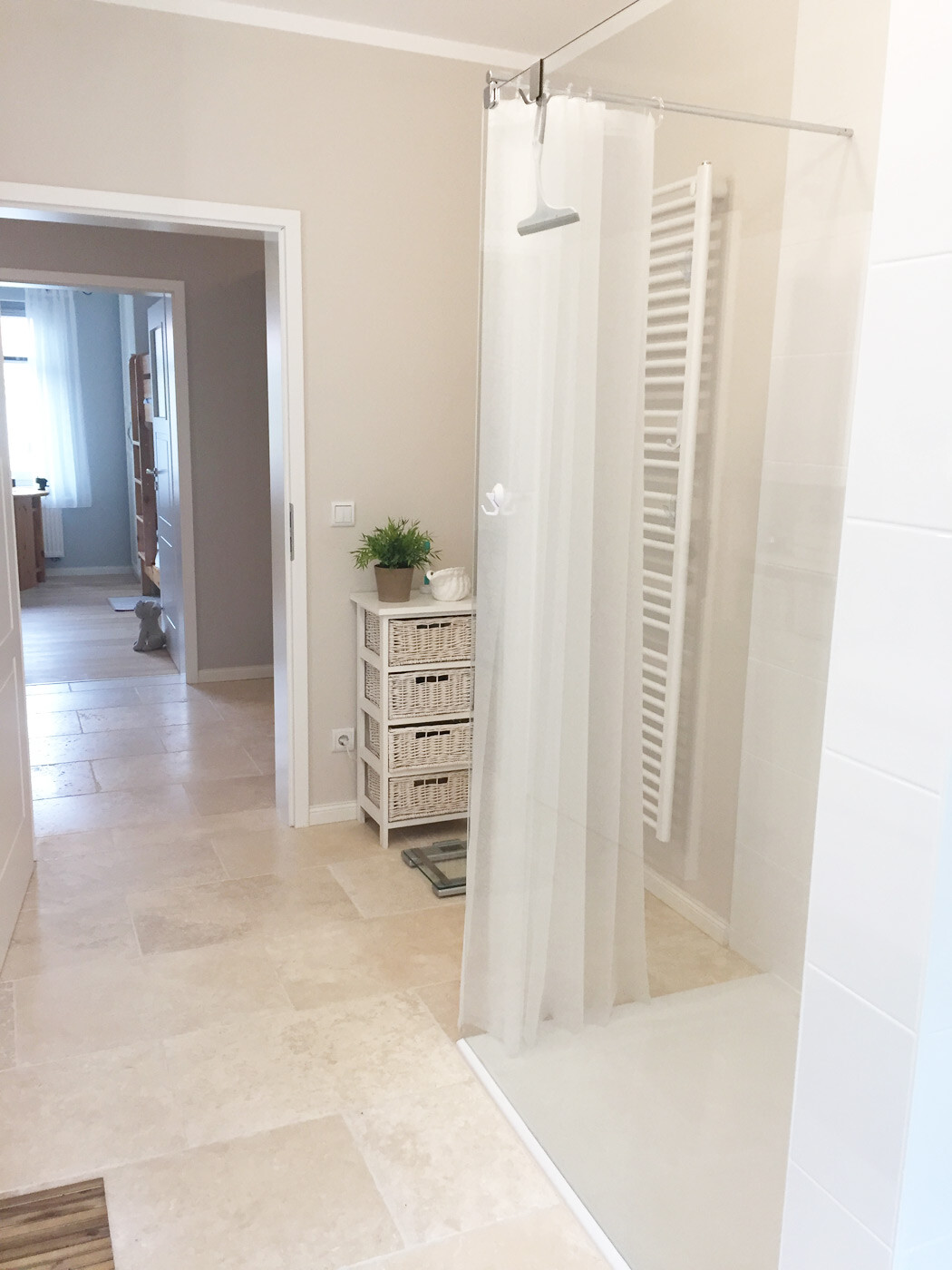 Badezimmer mit Duschabtrennung, Vorhang, Heizkörper, Korbkommode und Pflanze.