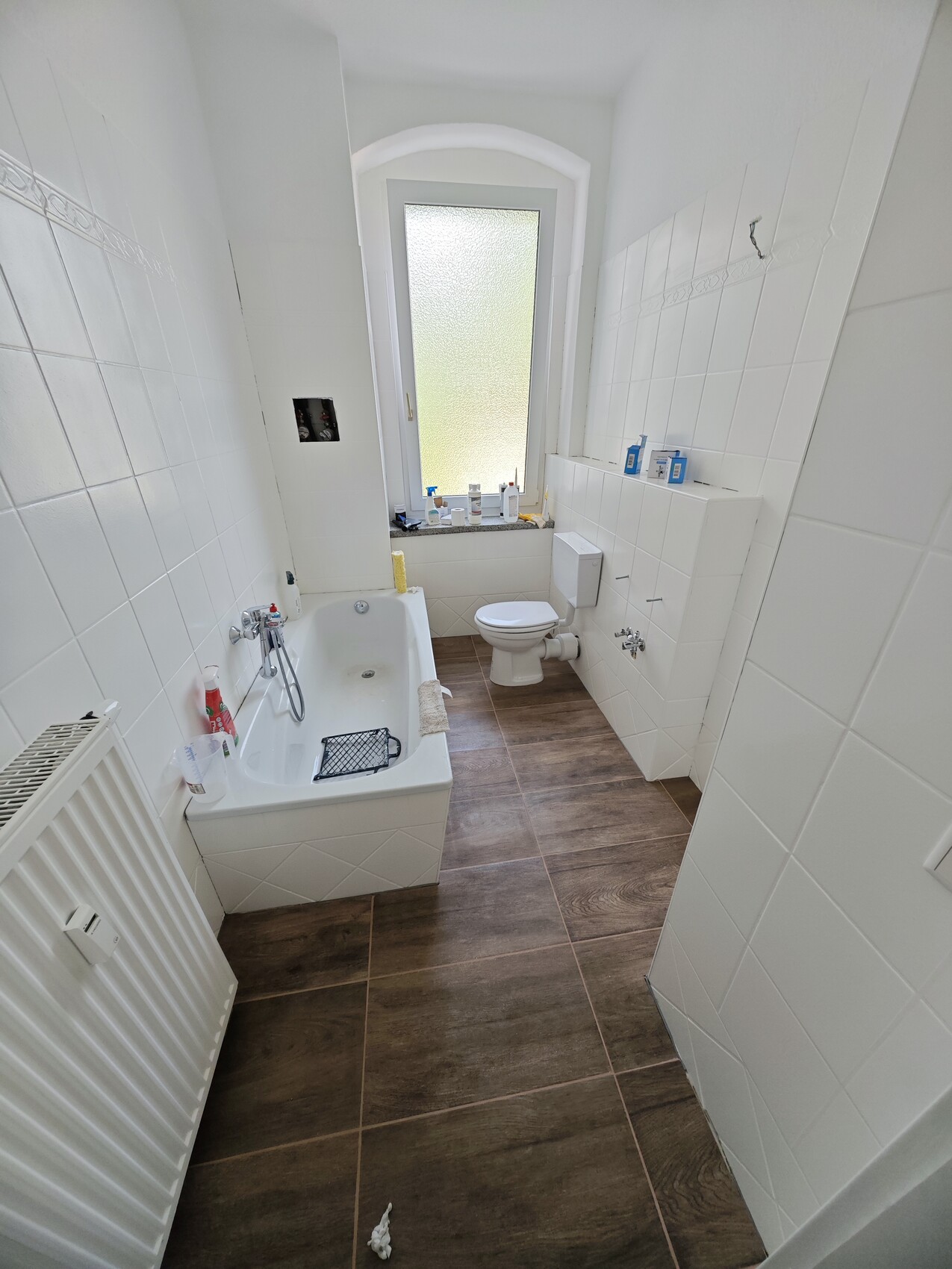 Helles Badezimmer mit weißer Fliesenwand, Badewanne links, Fenster hinten, Toilette, brauner Boden.