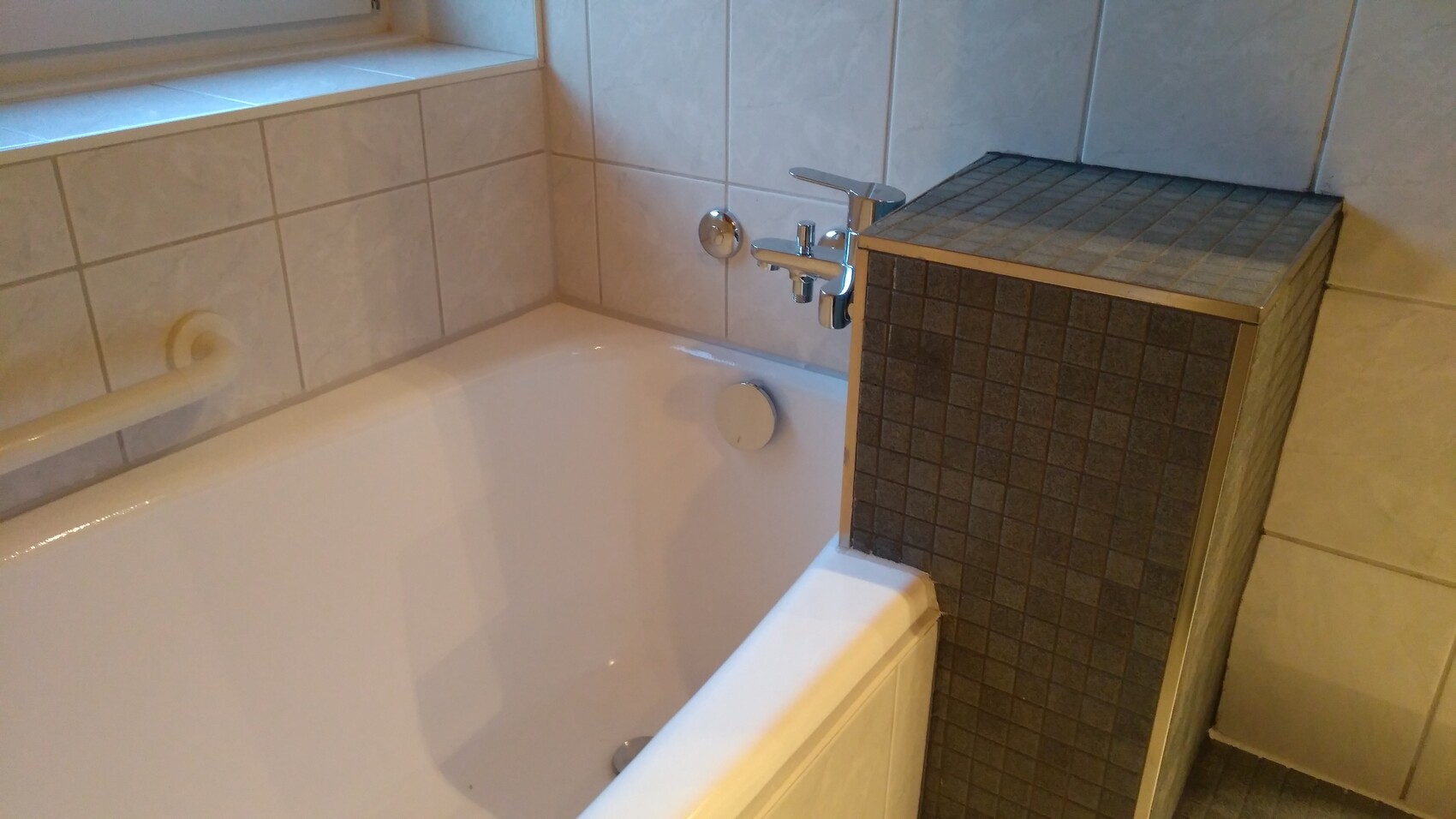 Badezimmer-Eckbadewanne mit Chrom-Armatur neben einem mosaikverkleideten Block.