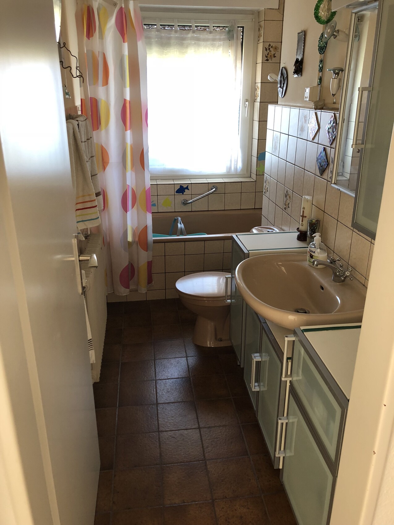 Kleines Badezimmer mit Waschbecken, Toilette, Badewanne und bunten Duschvorhang vor dem Fenster.