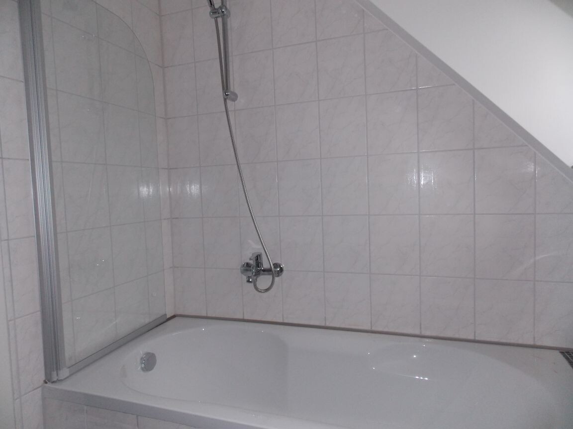 Weiße Badewanne mit Dusche, Handbrause und Glasabtrennung vor Fliesenwand.