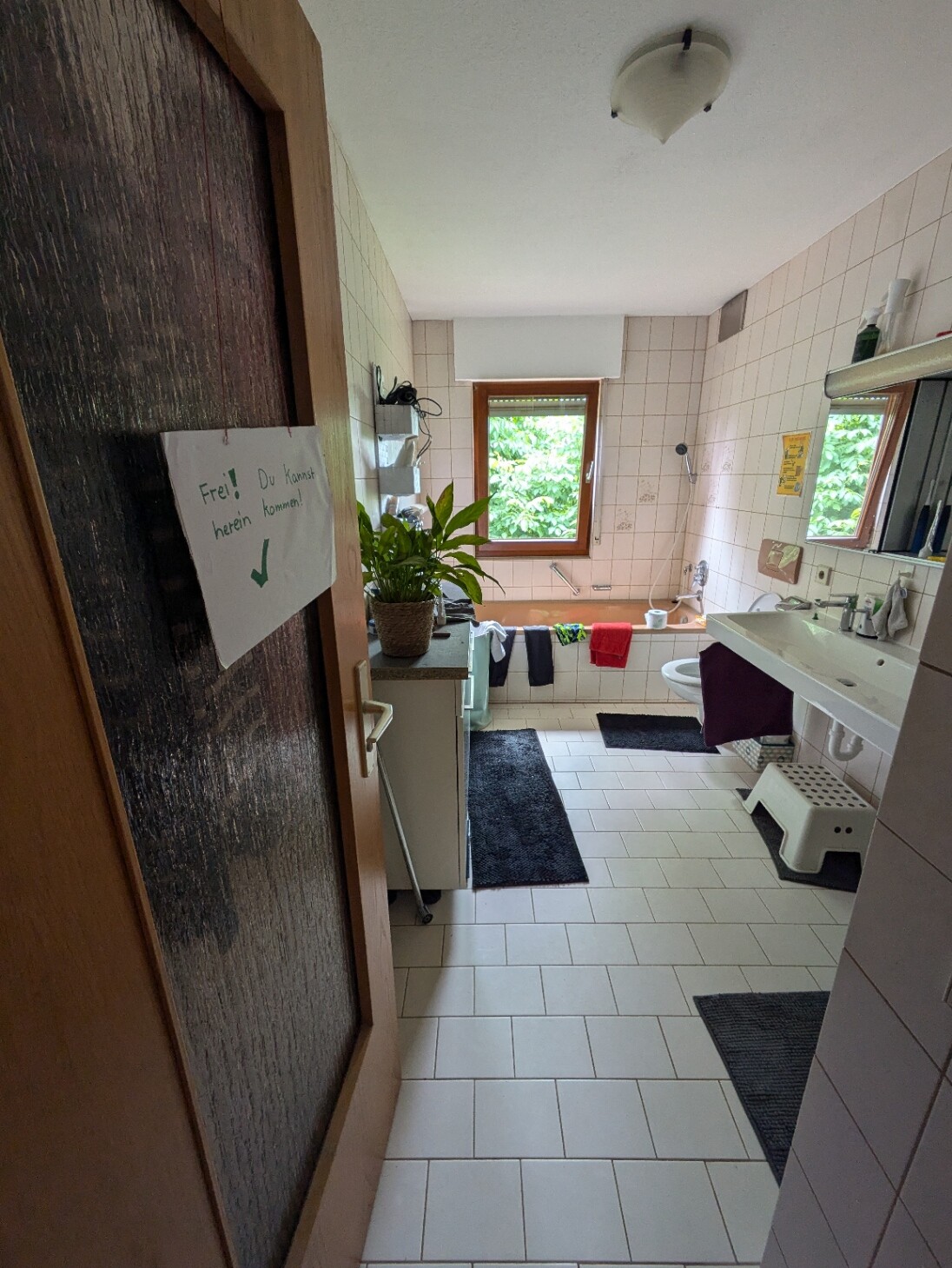Badezimmer mit Fenster, Waschbecken, Badewanne und Pflanze; Türschild 'Frei! Du kommst herein'.