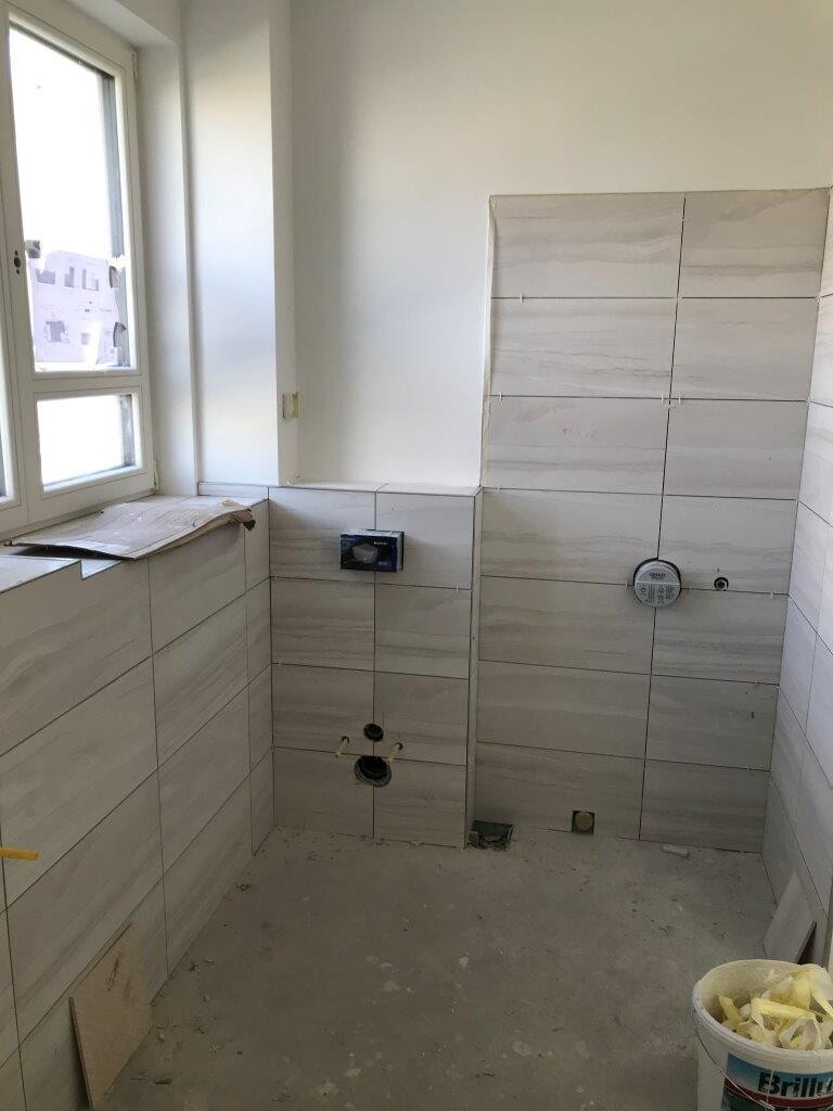 Badezimmer im Rohbau, hellgraue Fliesen, Fenster links, sichtbare Installationen, Eimer rechts.