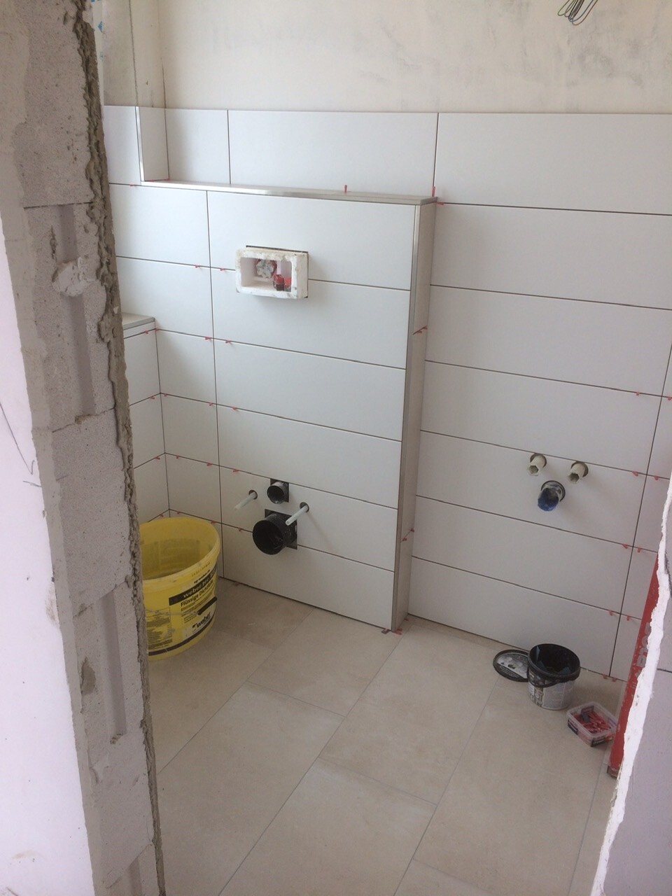 Rohbau-Badezimmer mit weißen Fliesen, Rohranschlüssen und Eimer auf dem Boden.
