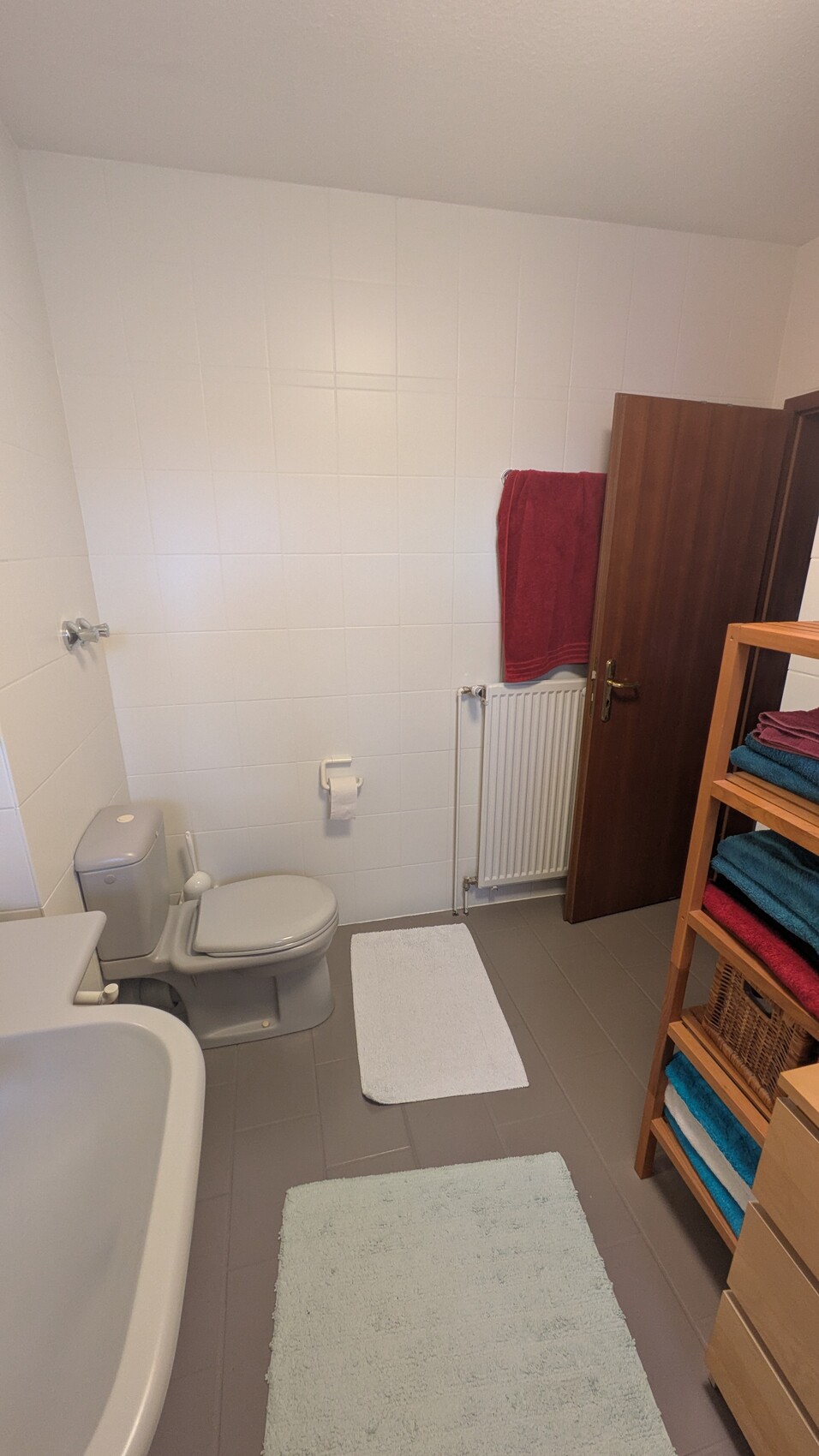 Helles Badezimmer mit Toilette, Waschbecken, Badewanne, Heizkörper und Regal mit Handtüchern.