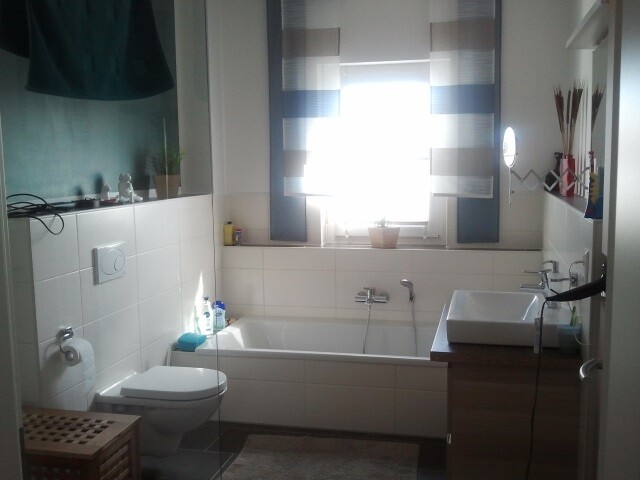 Helles Badezimmer mit Badewanne, Toilette, Waschbecken und Fenster mit Rollläden
