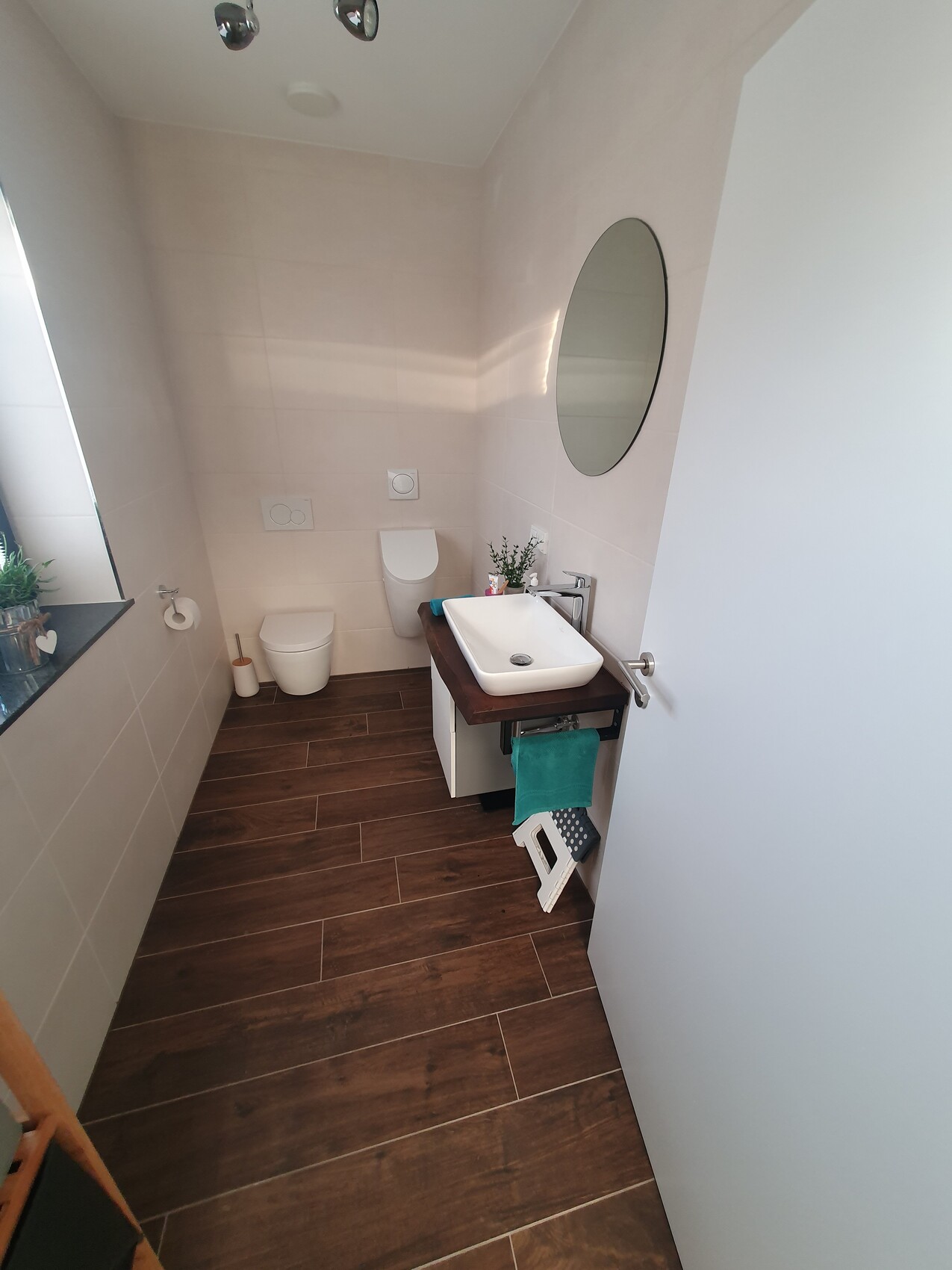 Kleines modernes Bad mit wandhängendem WC, Waschbecken auf Holzplatte, ovalem Spiegel.