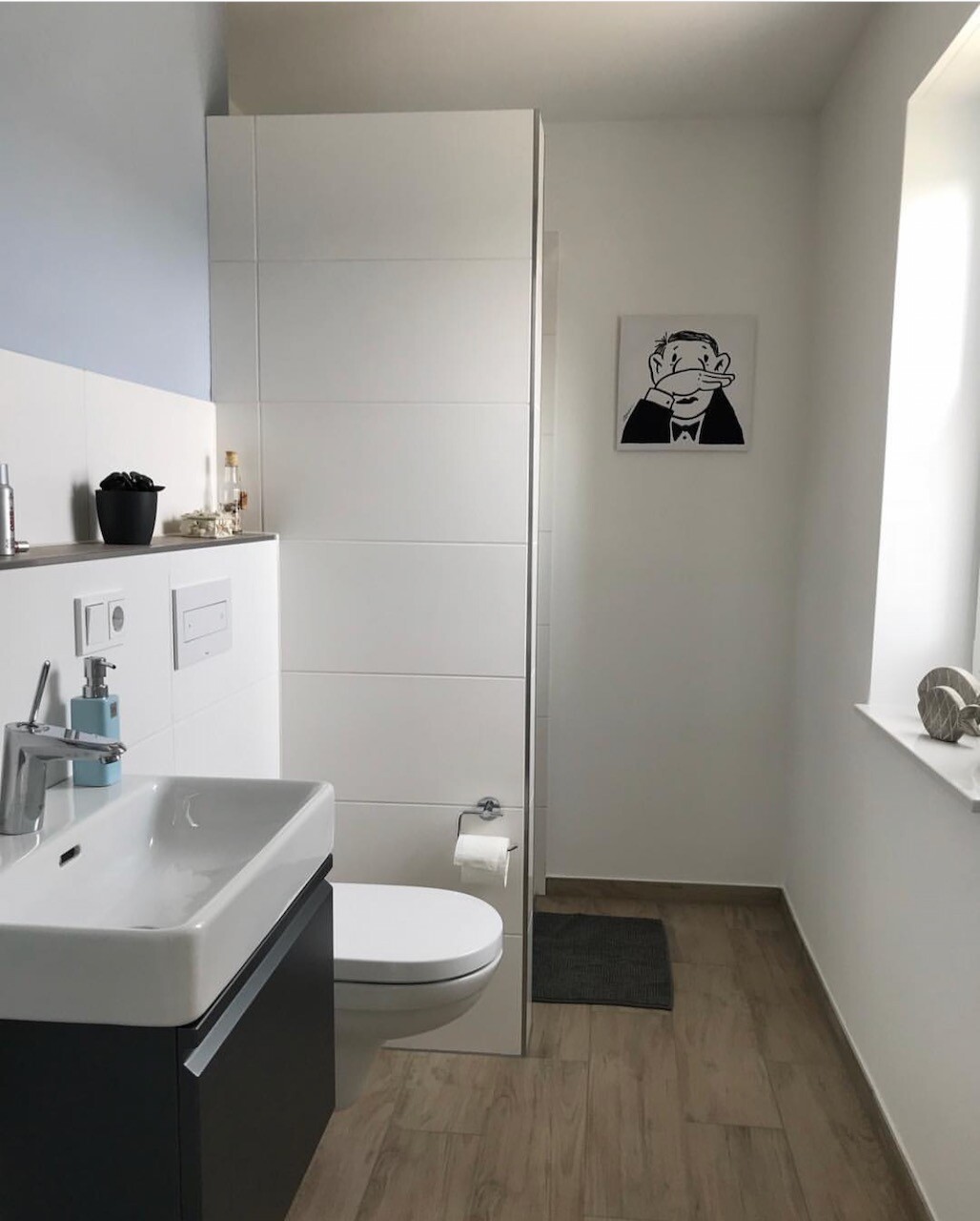 Modernes Badezimmer mit Waschbecken, Wandtoilette, weißen Fliesen, Unterschrank, Fenster und Poster.