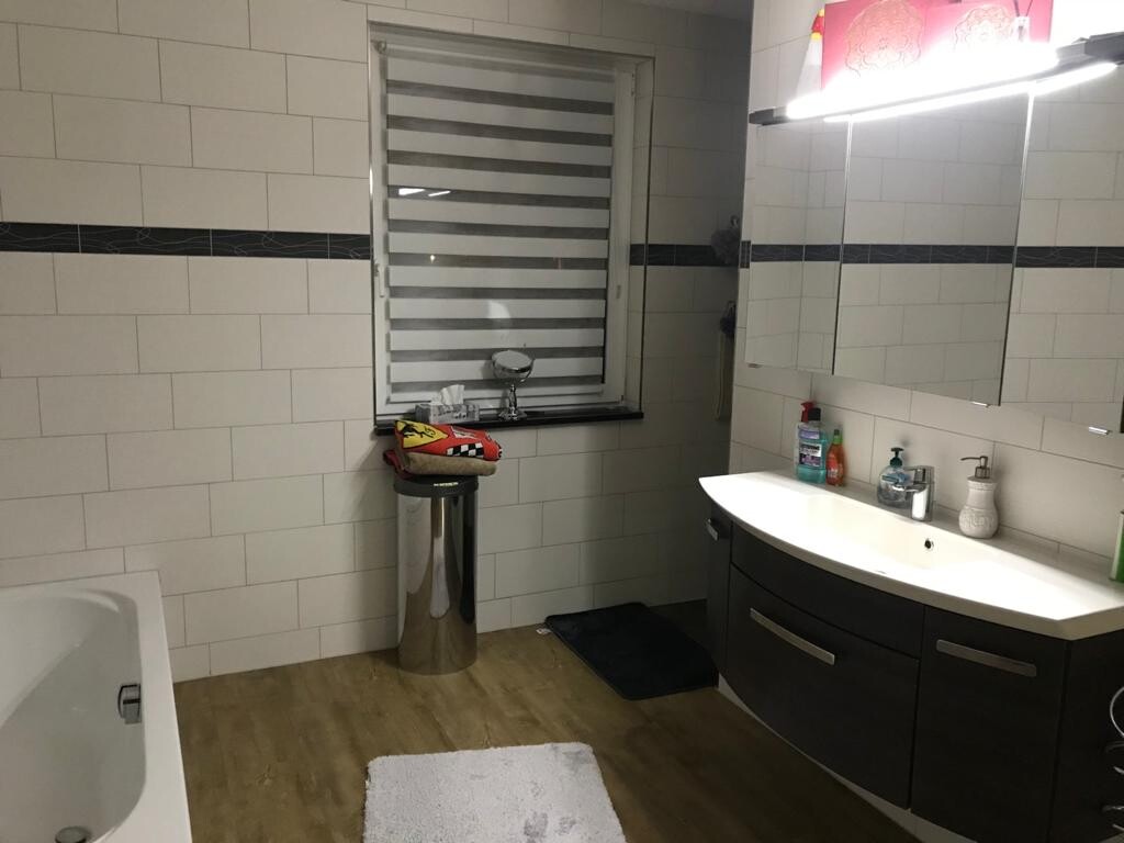 Modernes Badezimmer: weisse Fliesen, dunkler Waschtisch, Spiegelschrank, Fenster mit Jalousie.