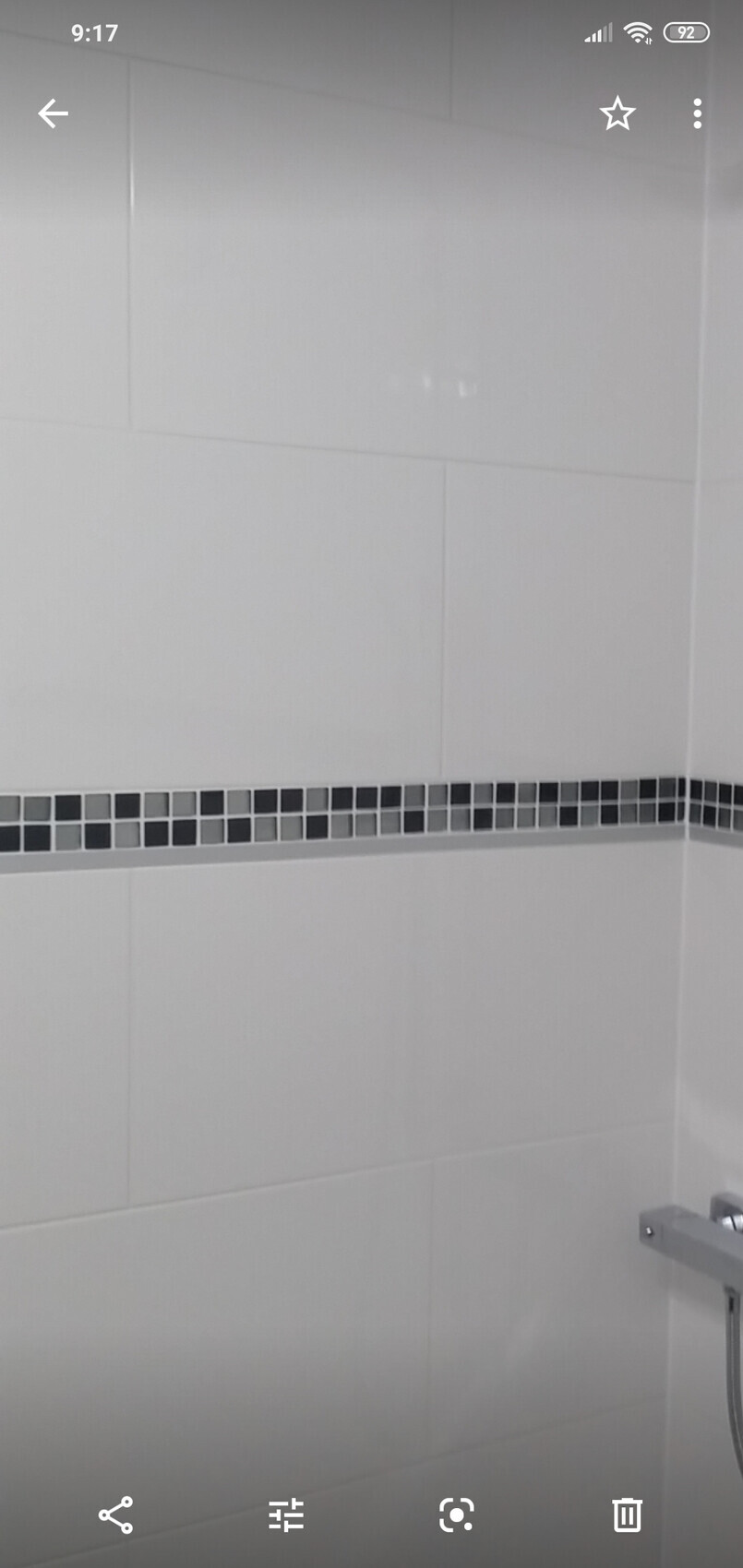 Weiße Badezimmerfliesen mit schwarzer Mosaikbordüre; Duscharmatur rechts unten.