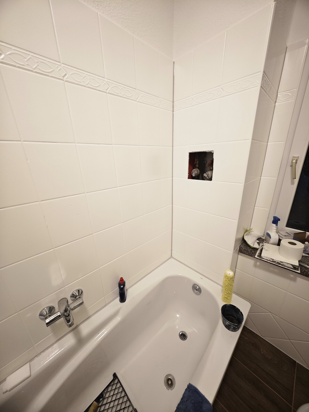 Weiß gefliestes Bad mit Badewanne, offenes Wandloch mit Rohren, Reinigungsmittel-Flasche am Wannenrand.
