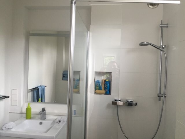Badezimmer mit Glasdusche, Spiegel, Waschbecken, Seifenspender, Handtuch und Regal mit Pflegeprodukten
