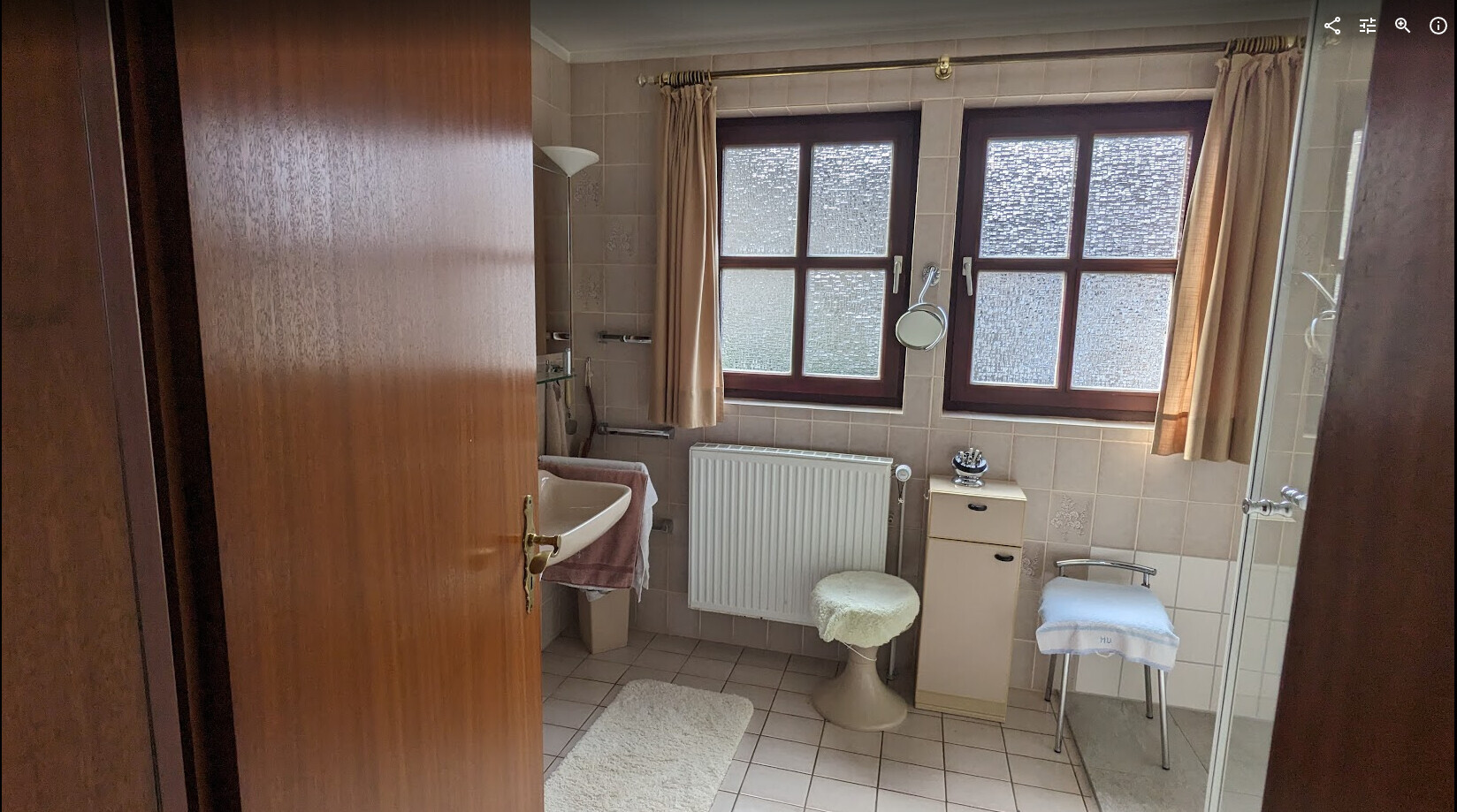 Badezimmer mit Waschbecken, Heizkörper, zwei Fenster, Stuhl und Duschkabine.